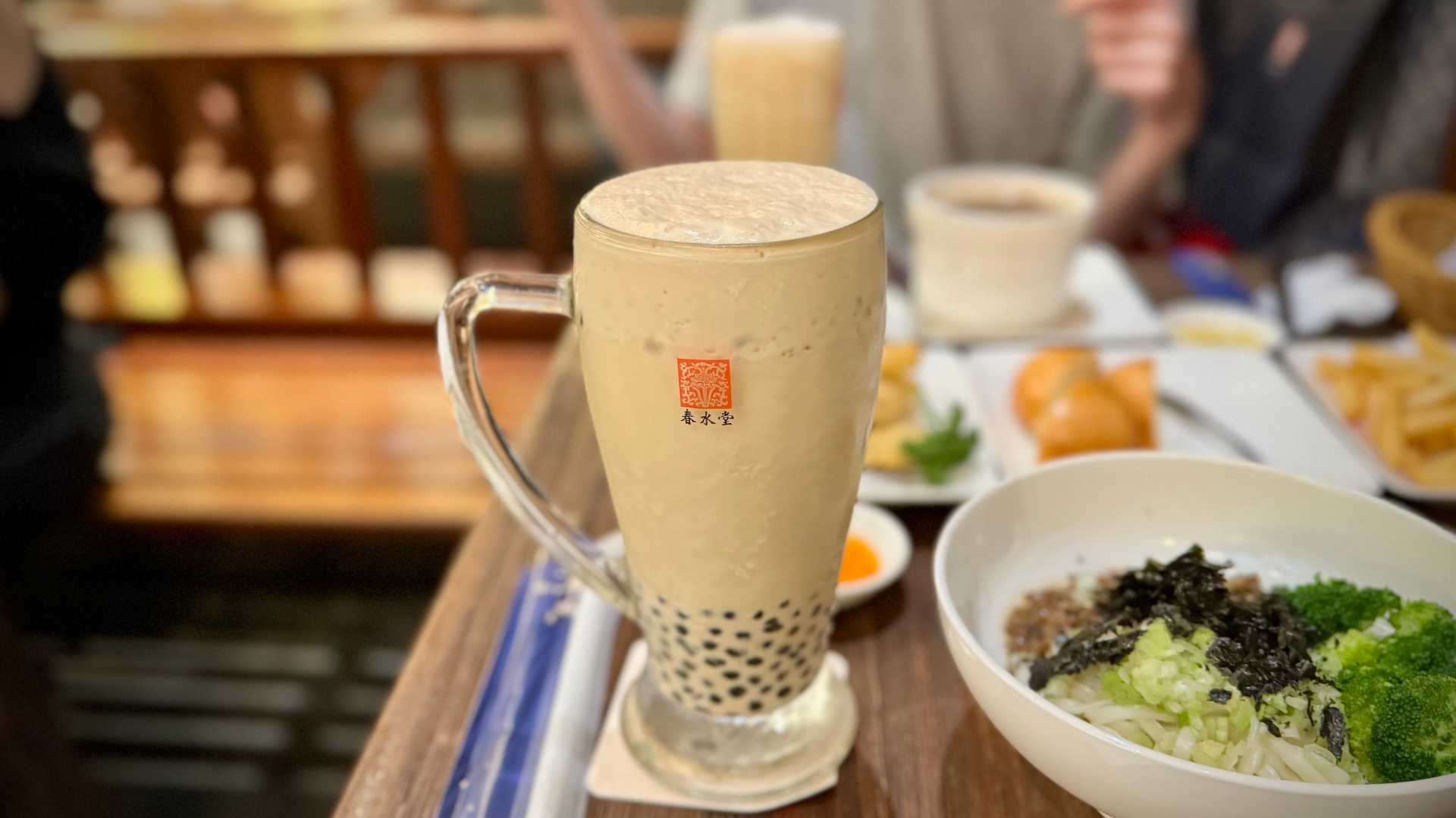 A large glass cup of iced milk bubble tea, on a table alongside various small dishes such as a bowl of noodles and vegetables, and some bread.