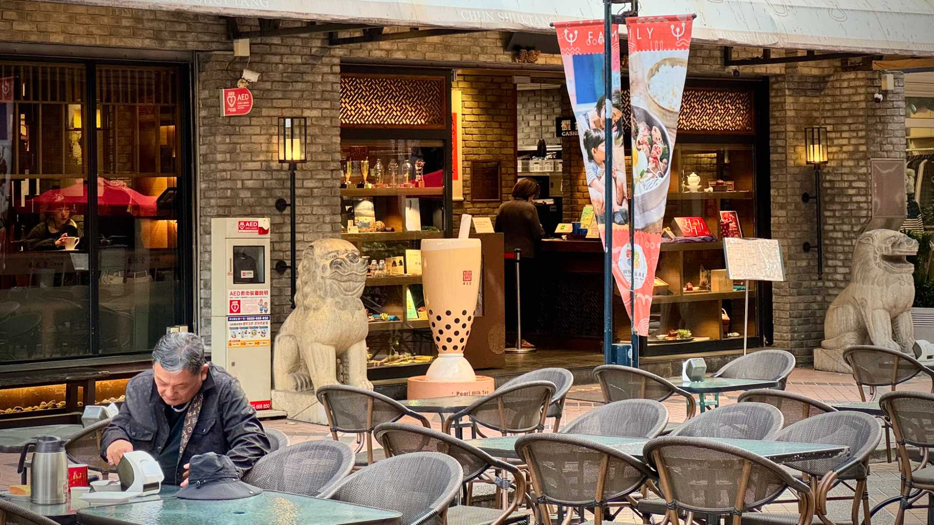 An elderly man sits alone drinking tea at one of many outdoor tables. Behind him is the entrance to a Chun Shui Tang teahouse, with stone lions either side of the door, plsu a 1.5 meter-tall sculpture of a glass of bubble tea next to one of the lions.
