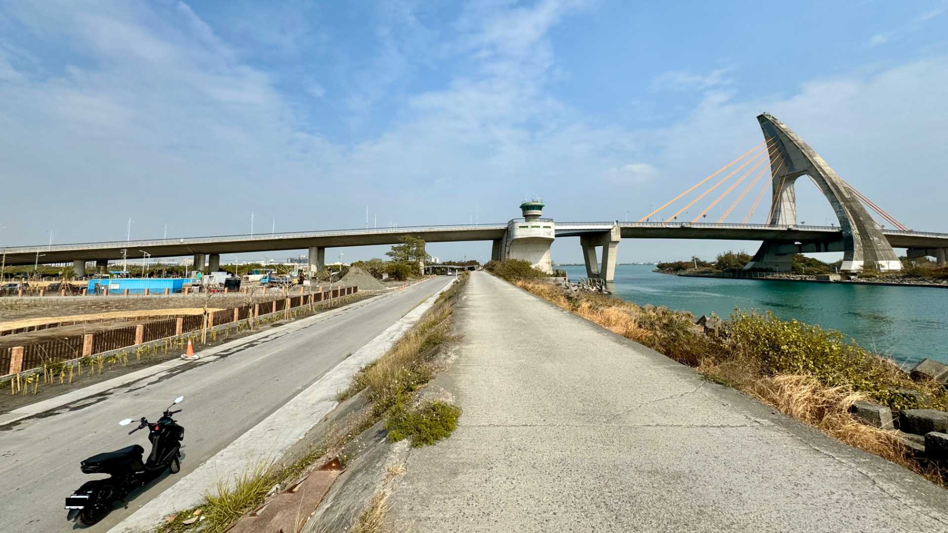 A scooter parked on a narrow road below Dapeng Bay Bridge. The bridge is in its lowered state.