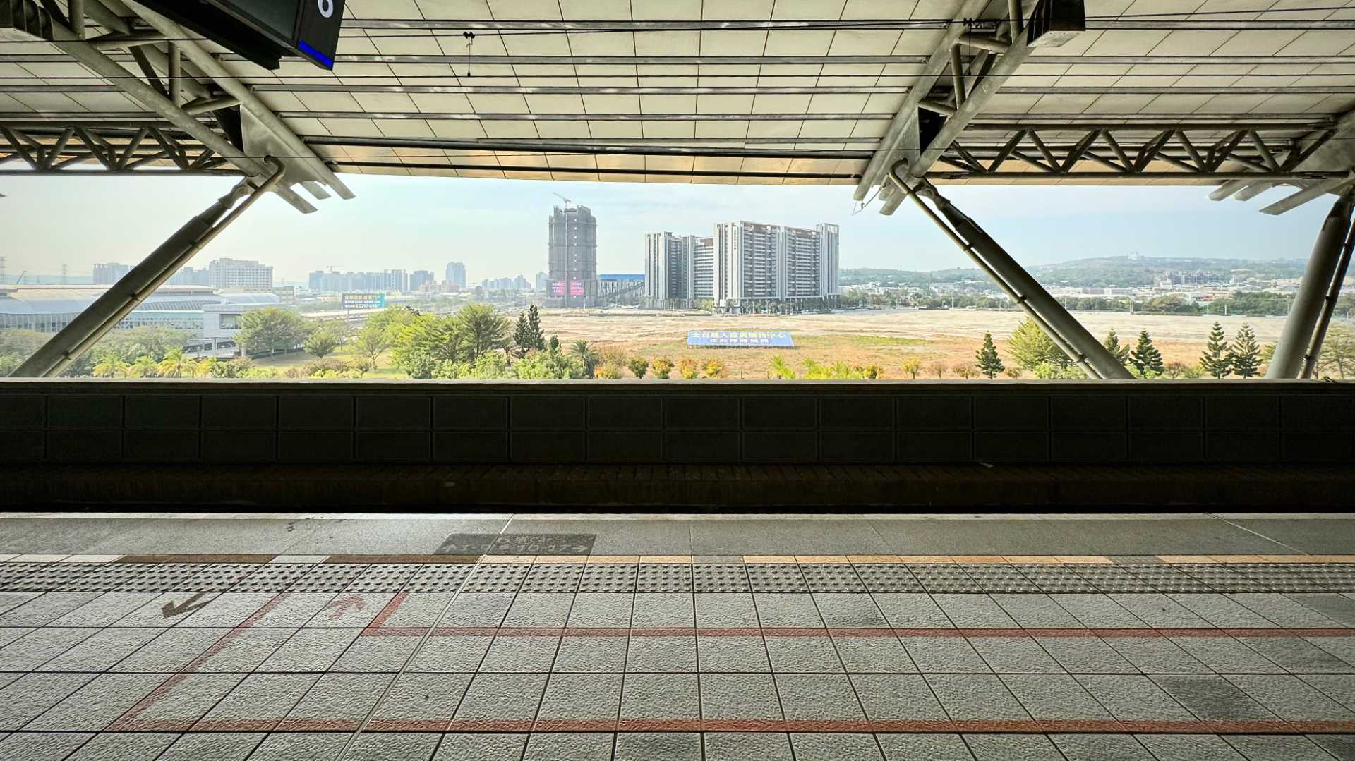 Daytime view looking from an elevated railway platform across some open fields, with city high rises and hills in the distance.