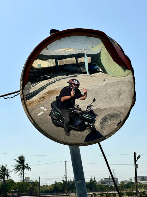 A distorted selfie taken in a convex roadside mirror. The photographer is sitting on his motorcycle and giving a peace sign with one hand.