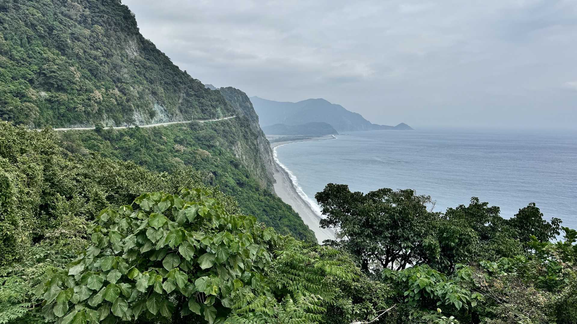 A wide-angle shot of a stunning piece of coastline, with jungle-clad mountains rising from the sea. A narrow road hugs the side of an extremely-steep cliff in the distance.