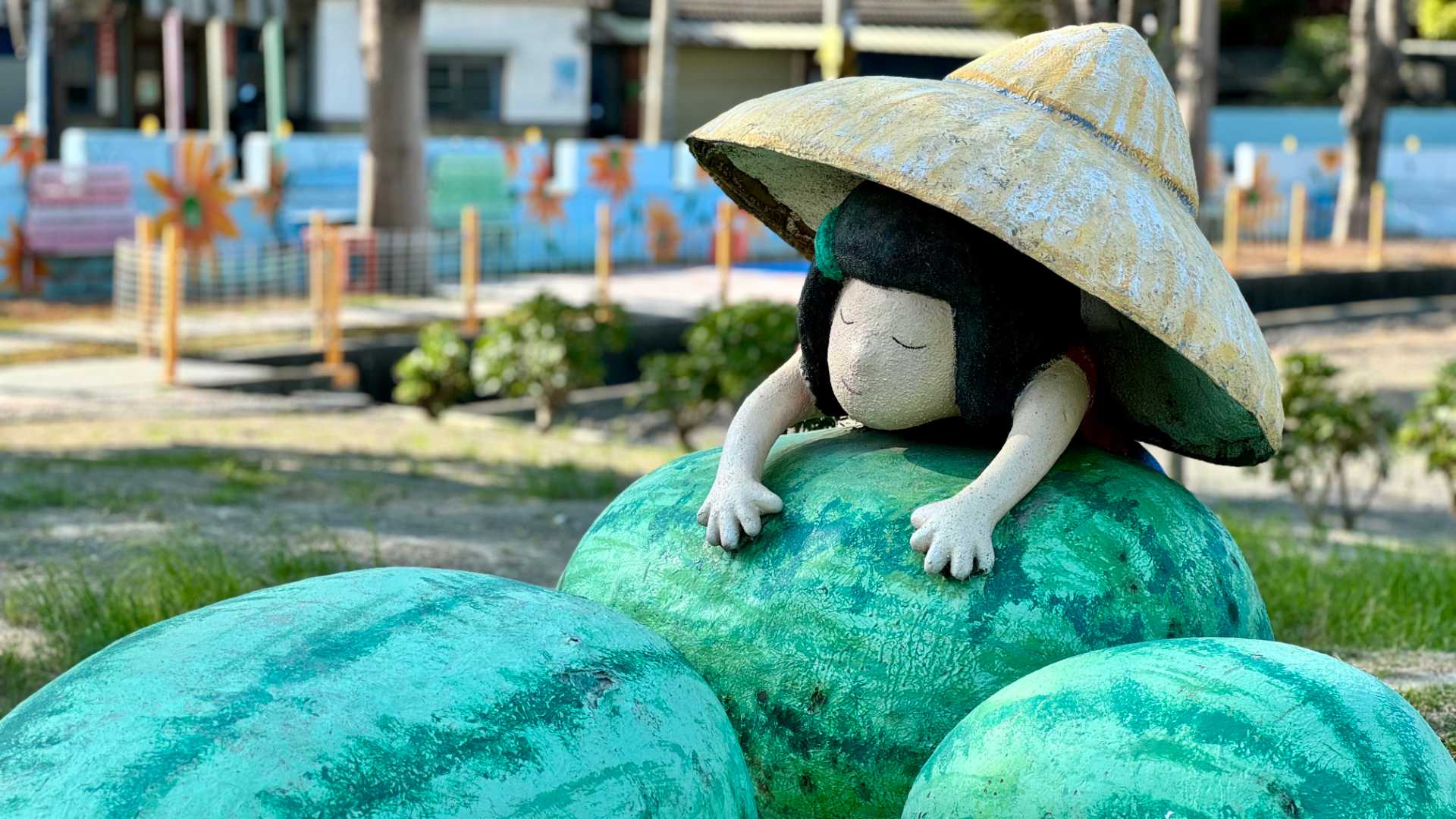 A sculpture of a girl, wearing an oversized triangular straw hat, leaning across some enormous melons.