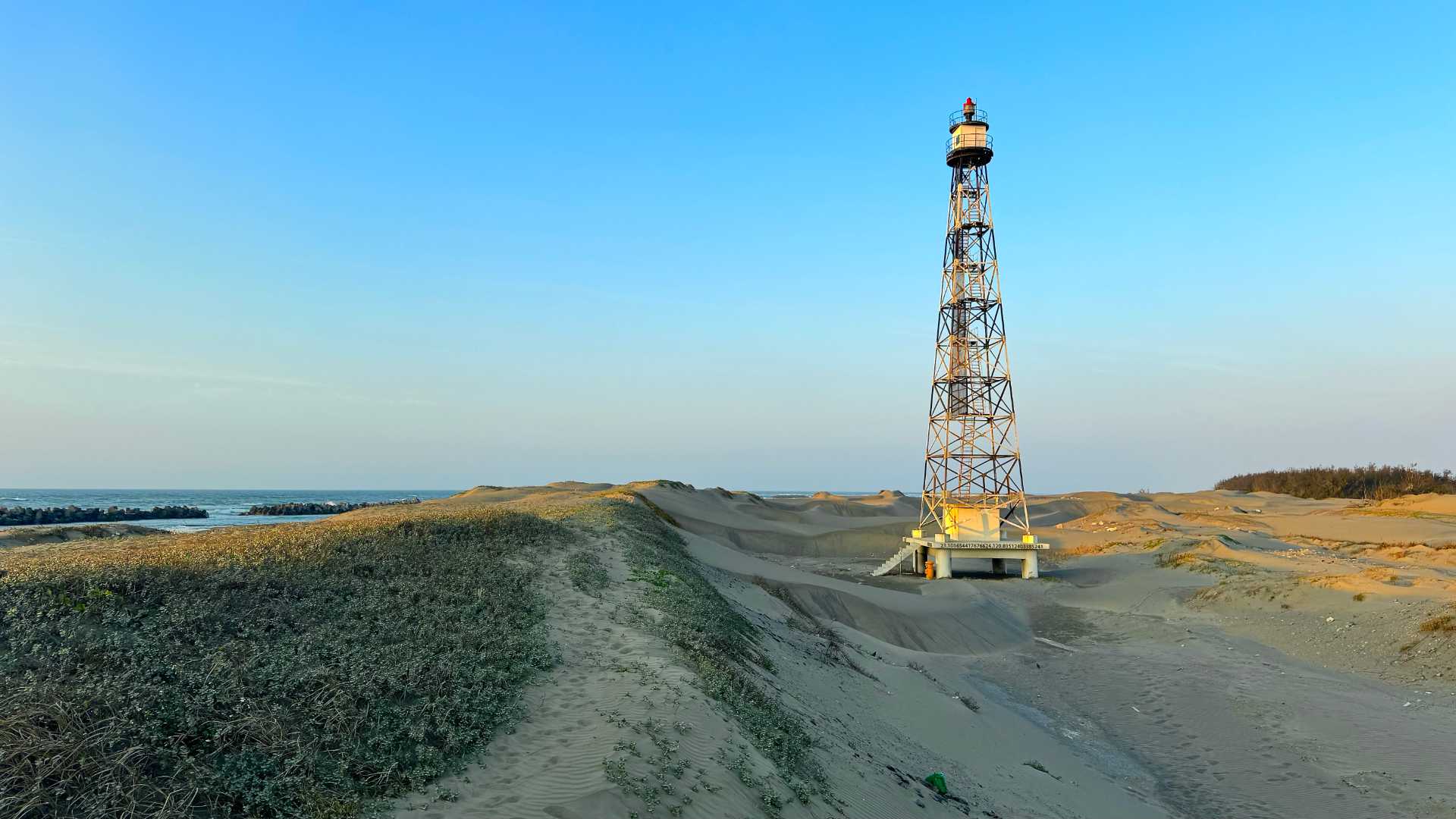 Guosheng Port Lighthouse, a skeletal metal structure rising from sand dunes, with the sea visible to the left.
