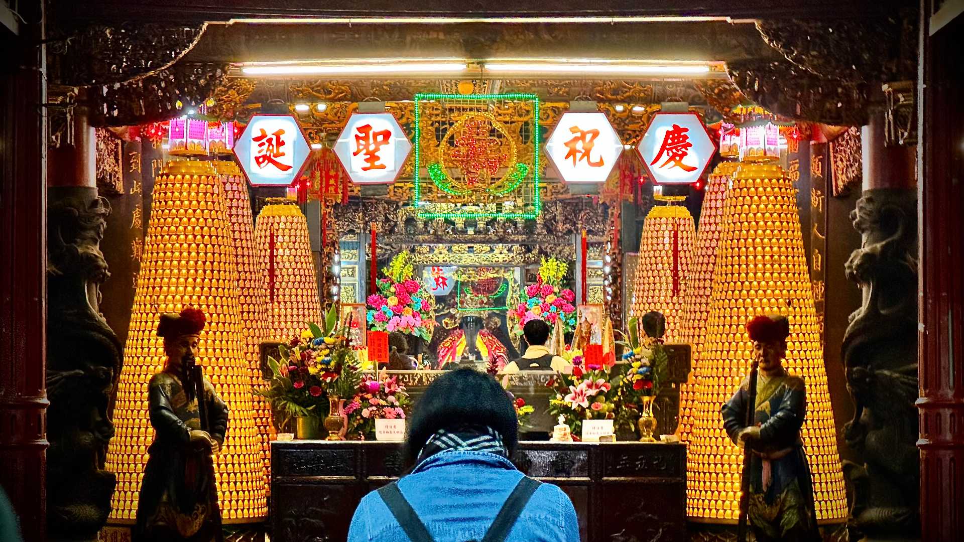 A woman praying inside Hsinchu Cheng Huang Temple. It is a richly-illuminated scene of flowers and a god on an alter, surrounded by thousands of electronic candles in a symmetrical arrangement.