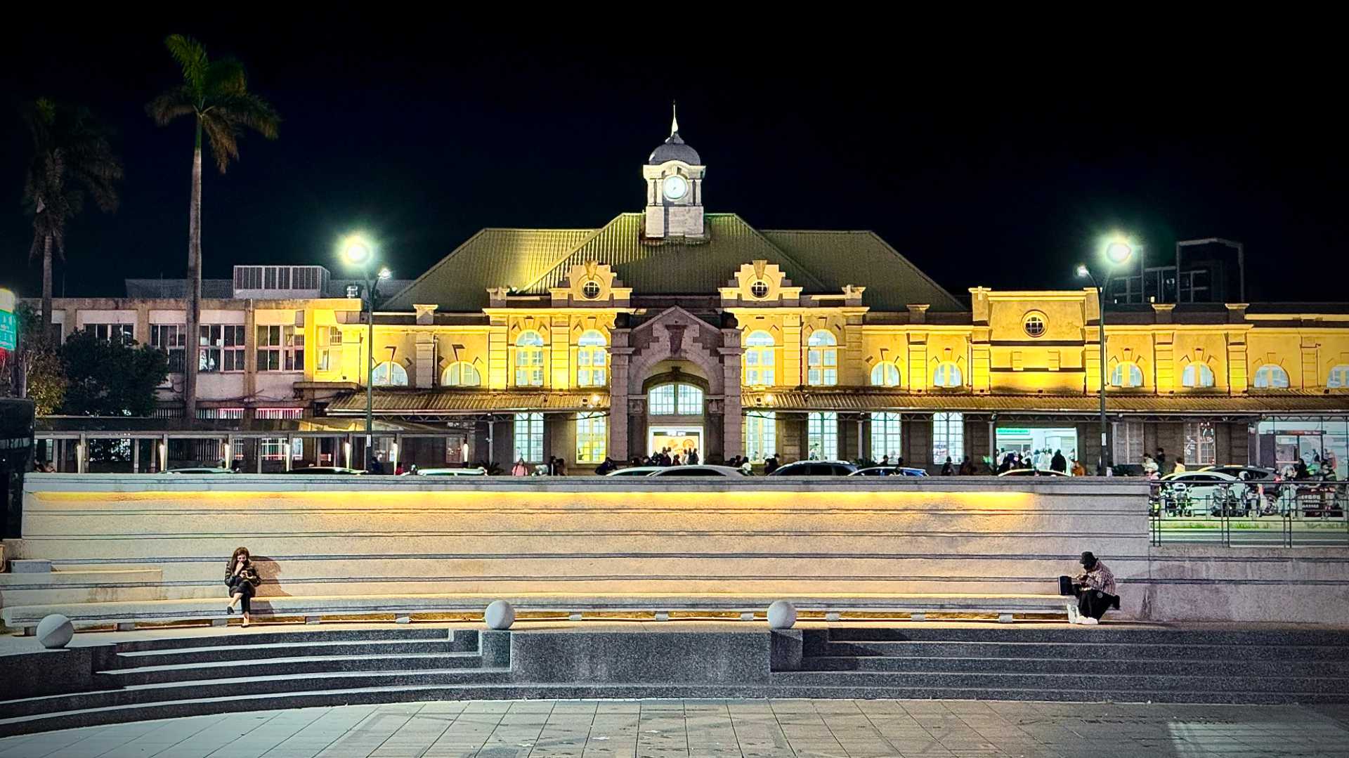 Wide-angle shot of Hsinchu Railway Station at night. Two people are sitting in a wide, shallow ampitheater in the foreground.