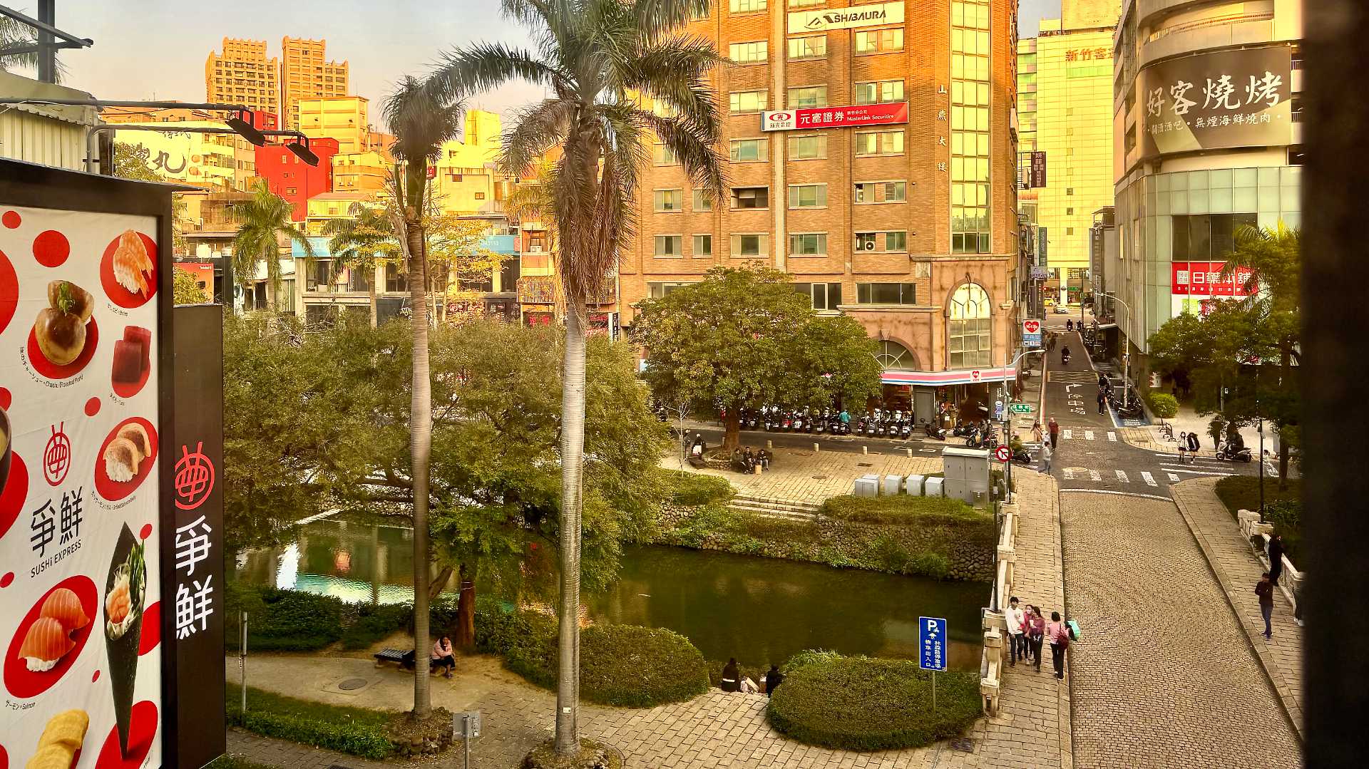 Photo of the center of Hsinchu city, showing a paved bridge across a canal lined with hedges and tall palm trees, with tall buildings in the distance.