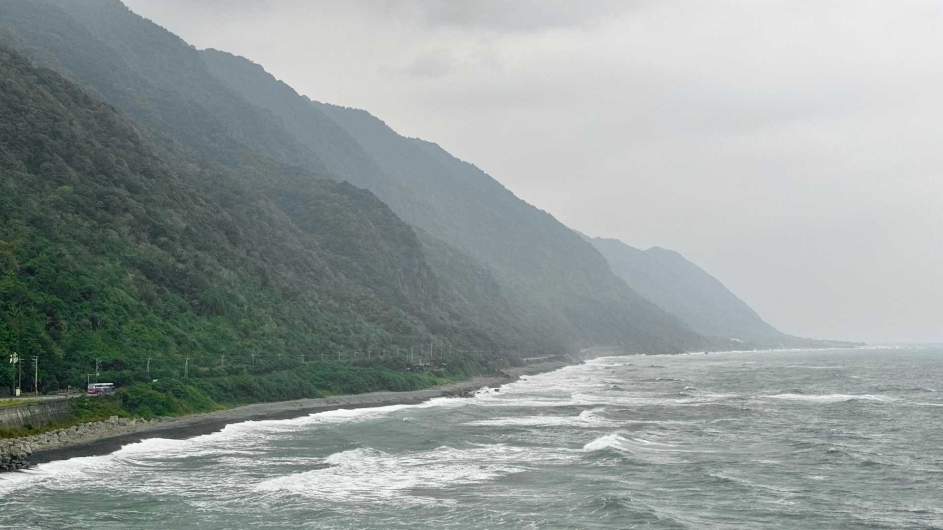 Large waves reaching the shore beneath forest-clad mountains. It is a misty day.