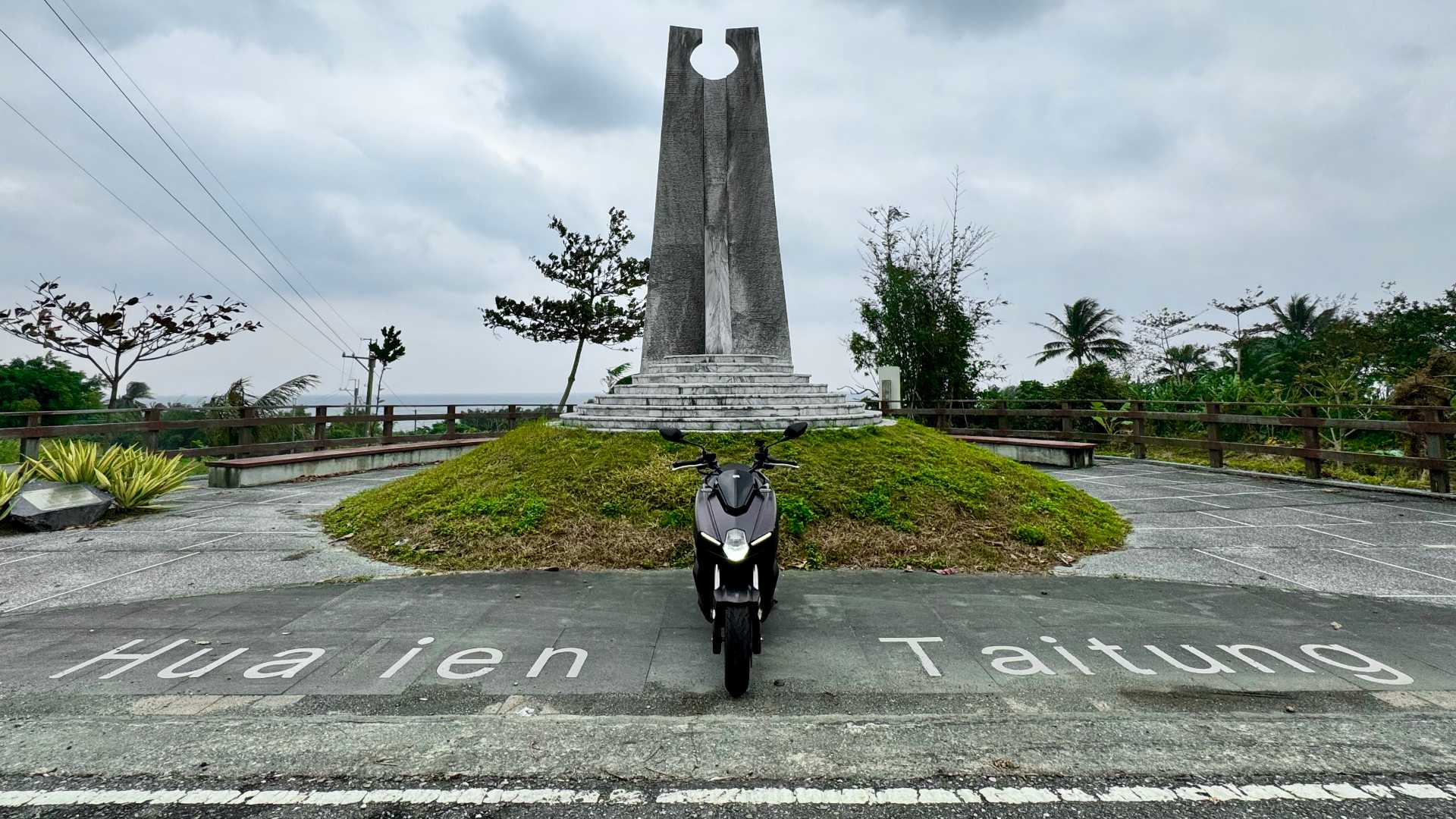 A tall, thin stone monument above the words ‘Hualien’ and ‘Taitung’ printed on the pavement. A motorcycle is parked between the two words, facing the camera with its lights on.