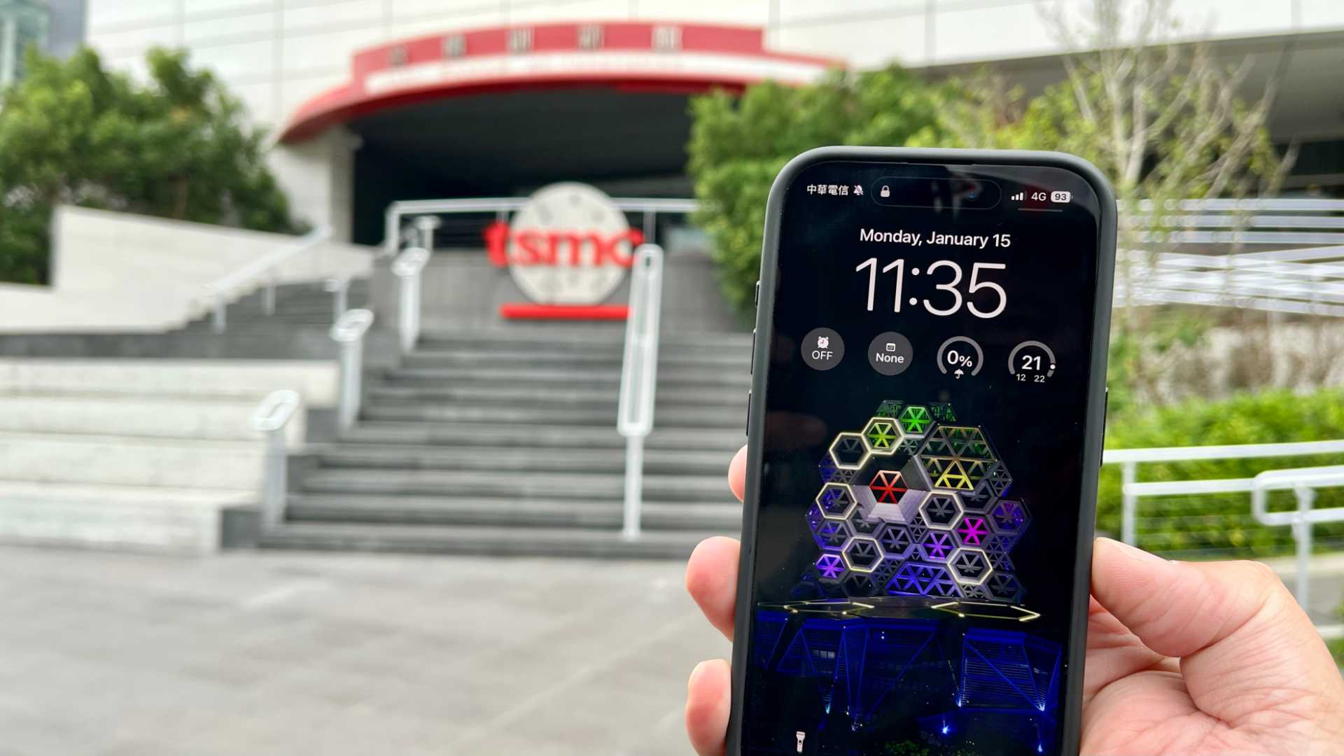An iPhone 15 Pro being held in front of the entrance to a TSMC building.