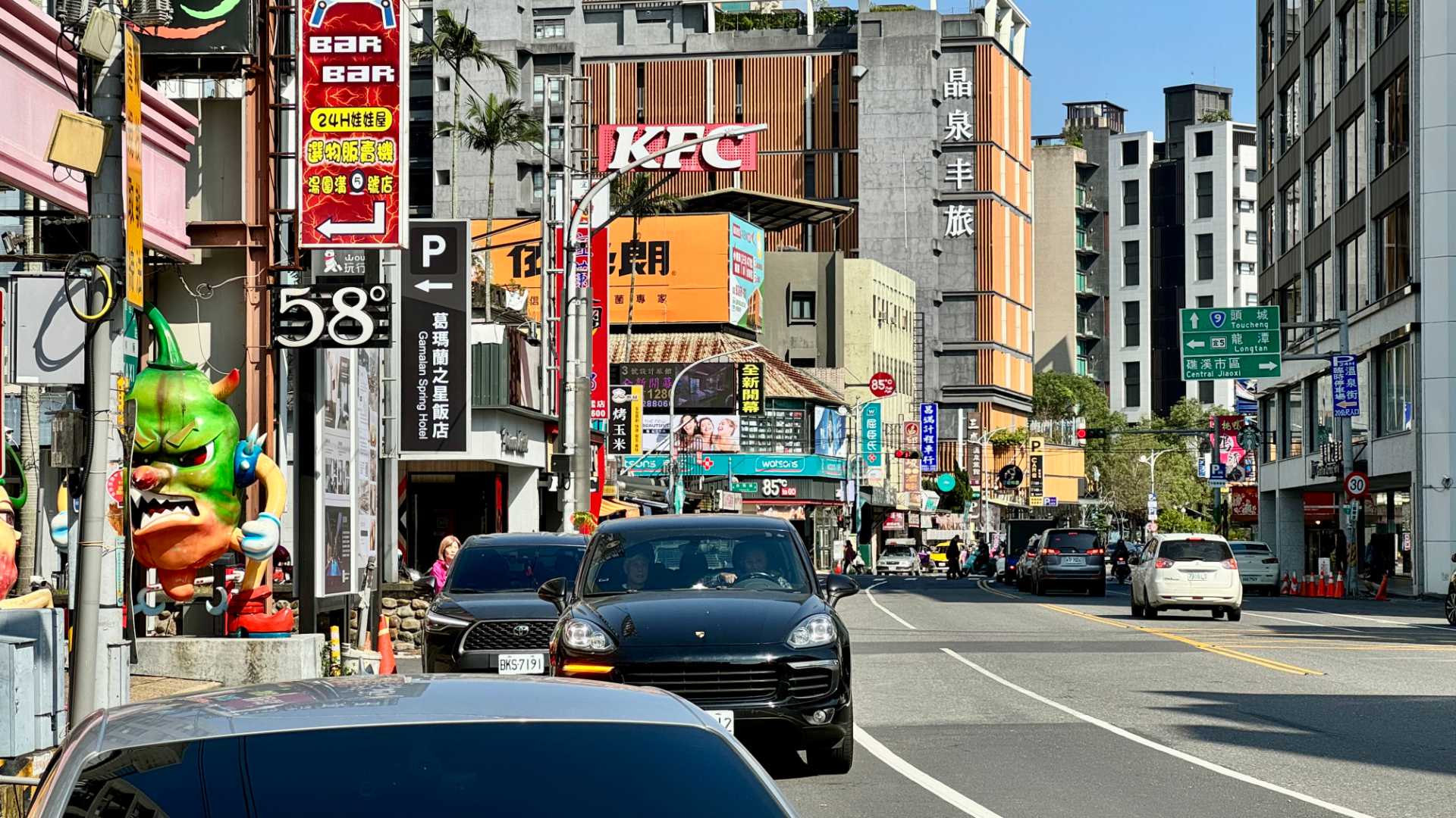 A bustling four-land road with shops and two-to-eight-story buildings on either side. It’s a sunny day.