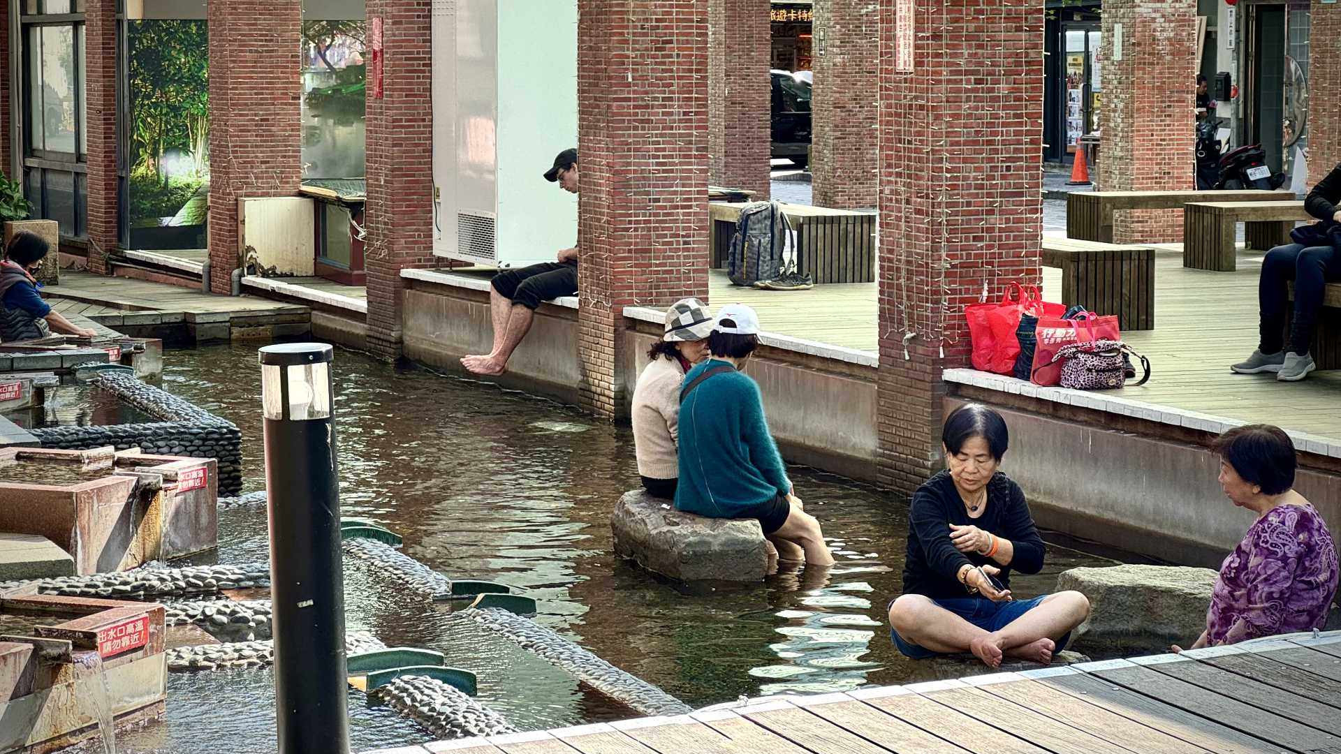 Six people sitting around a public hot-spring. Half of them have their legs in the hot water.