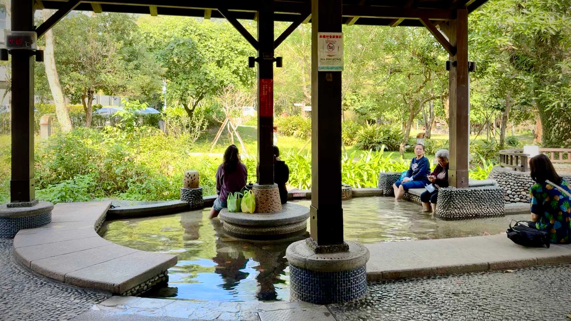 Five people sit around a public hot-spring, with their legs in the water. Beyond the hot-spring is a leafy green park.