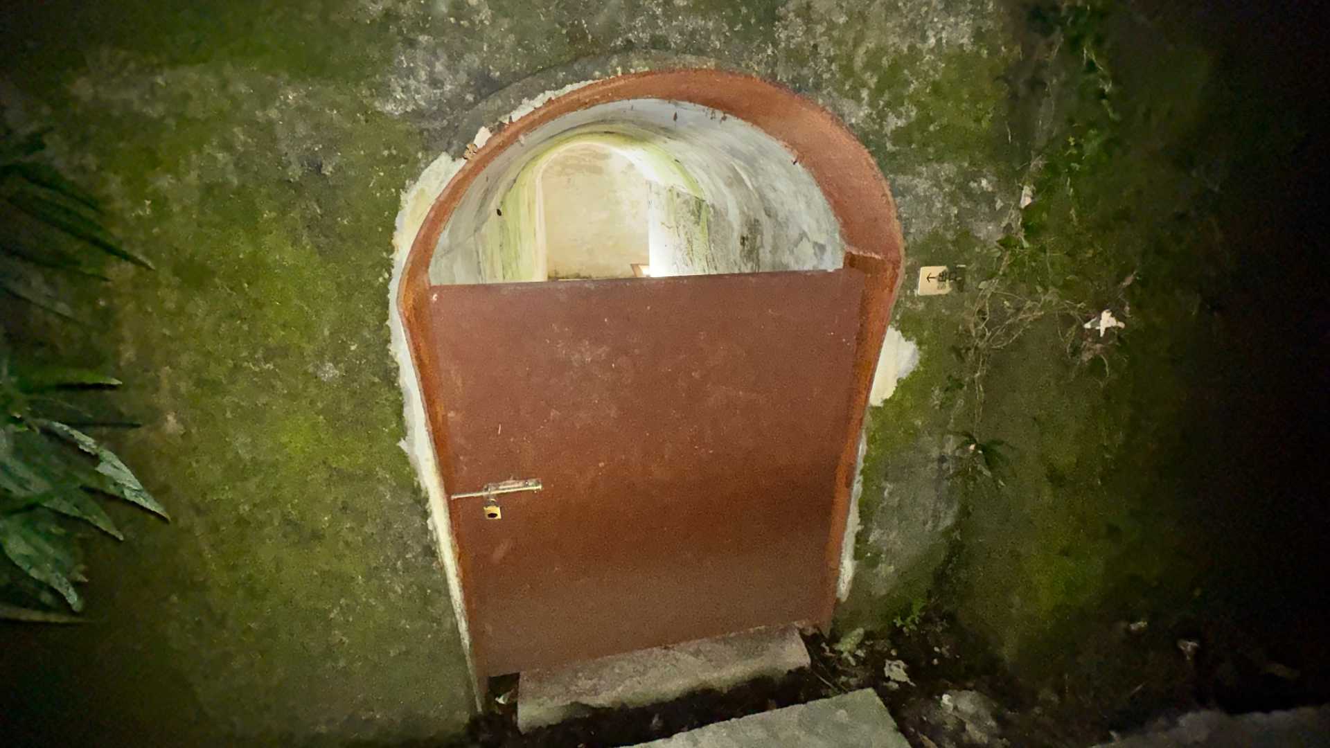 An arched entrance to a bomb shelter. A rusted metal door is locked, but the interior of the shelter can be seen over the top of the door. The walls are covered in green moss.