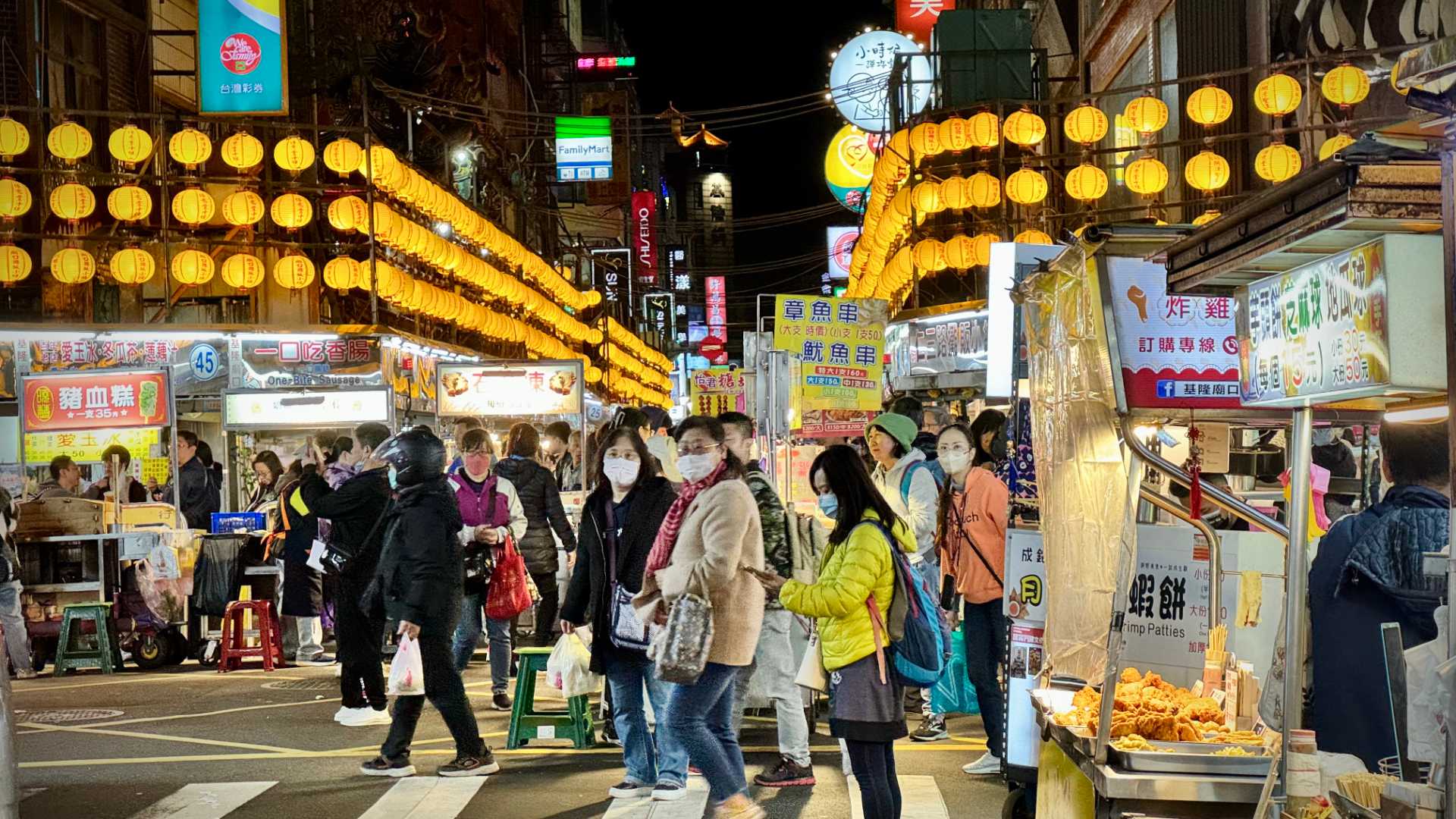 The entrance to a lively-looking, crowded night market in Keelung City. In the foreground, a stall is selling some kind of friend food. There are lanterns hanging above the street.