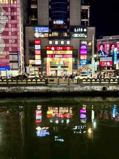 The lower few floors of a skyscraper reflected in a canal. The exterior walls of these lower floors are covered in illuminated signage for building tenants such as McDonalds, Muji, and ABC Mart. To the right of the building is a two-story-tall political billboard with a man and woman on it.