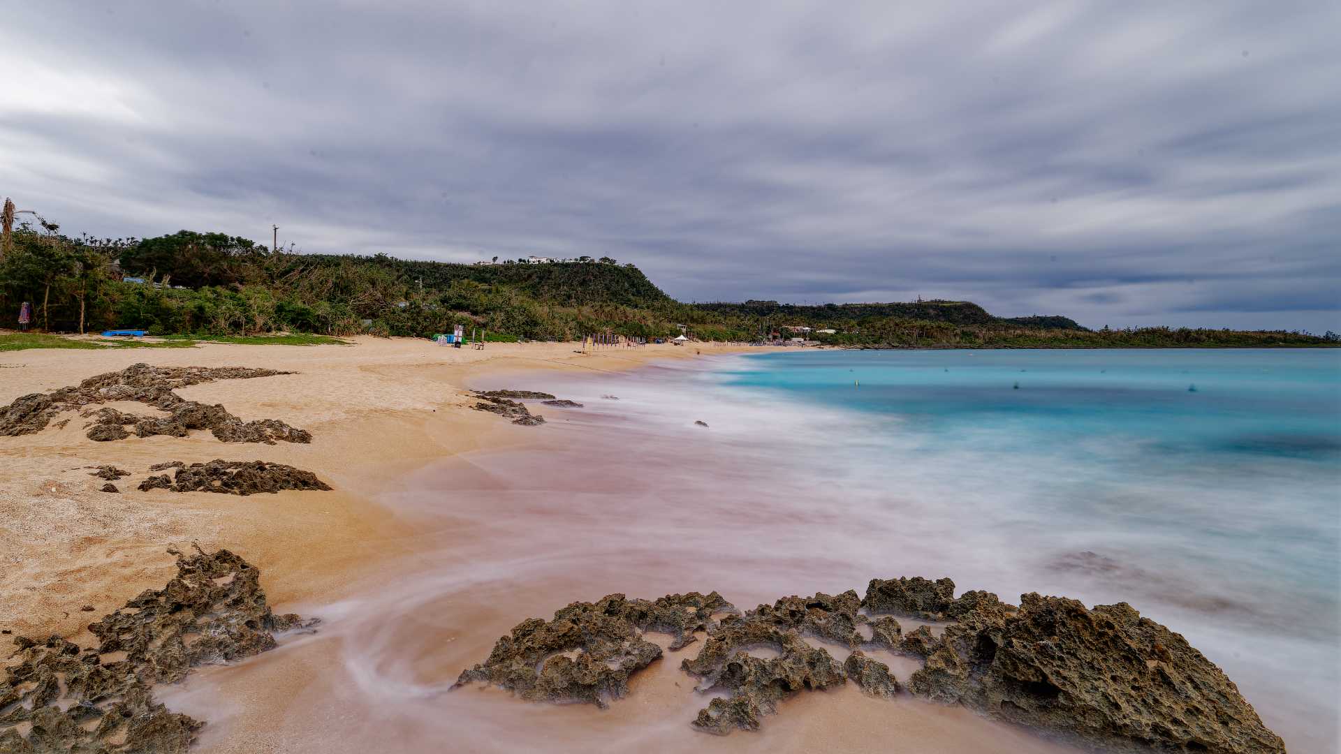 A long exposure of Kenting Baishawan Beach. The sand looks warm and golden, and the water is a vibrant, clear, azure blue. There are few people on the beach.