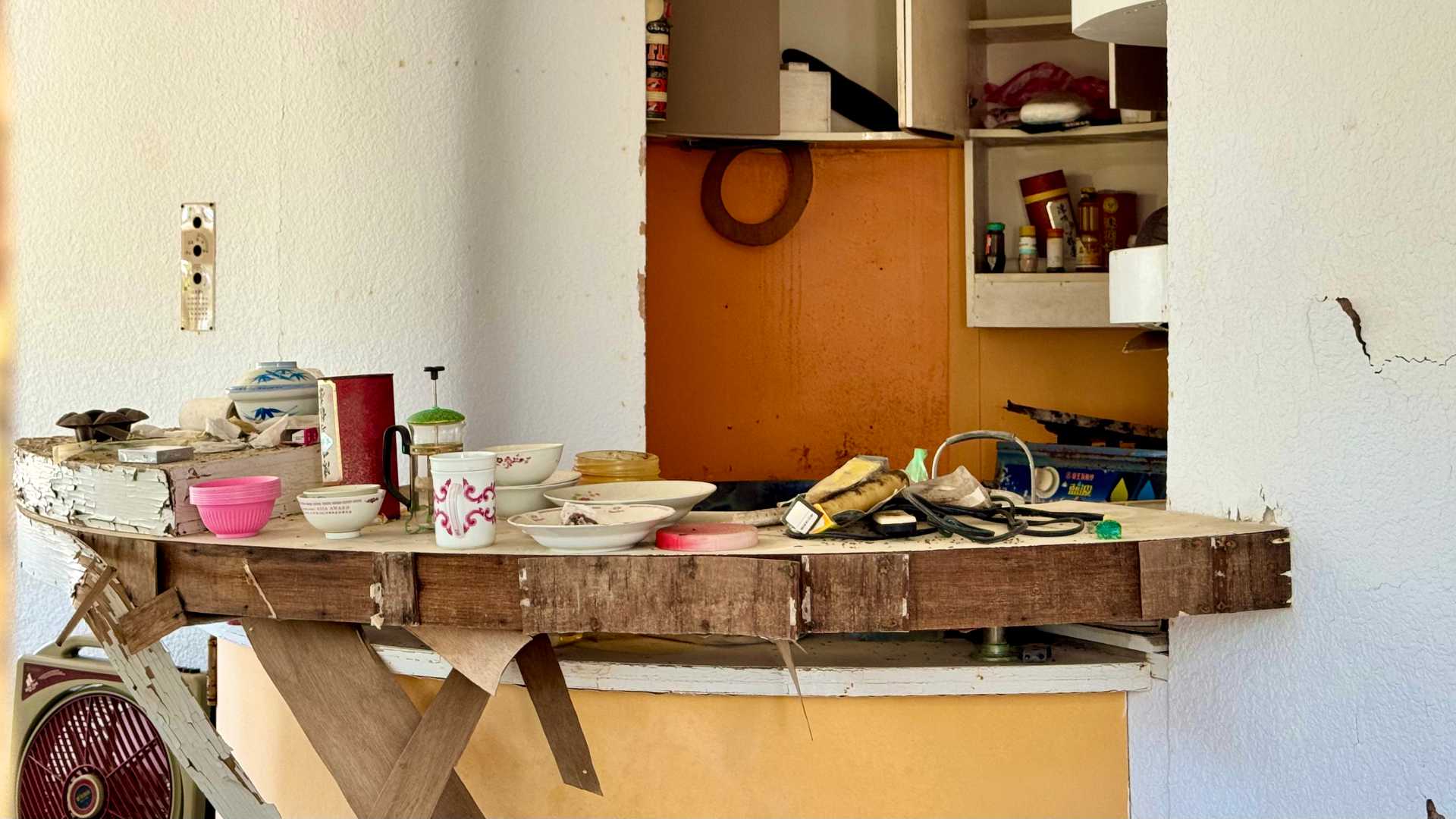 A partially-dismantled and broken kitchen, with plates, a coffee cup, and a coffee plunger on the counter.