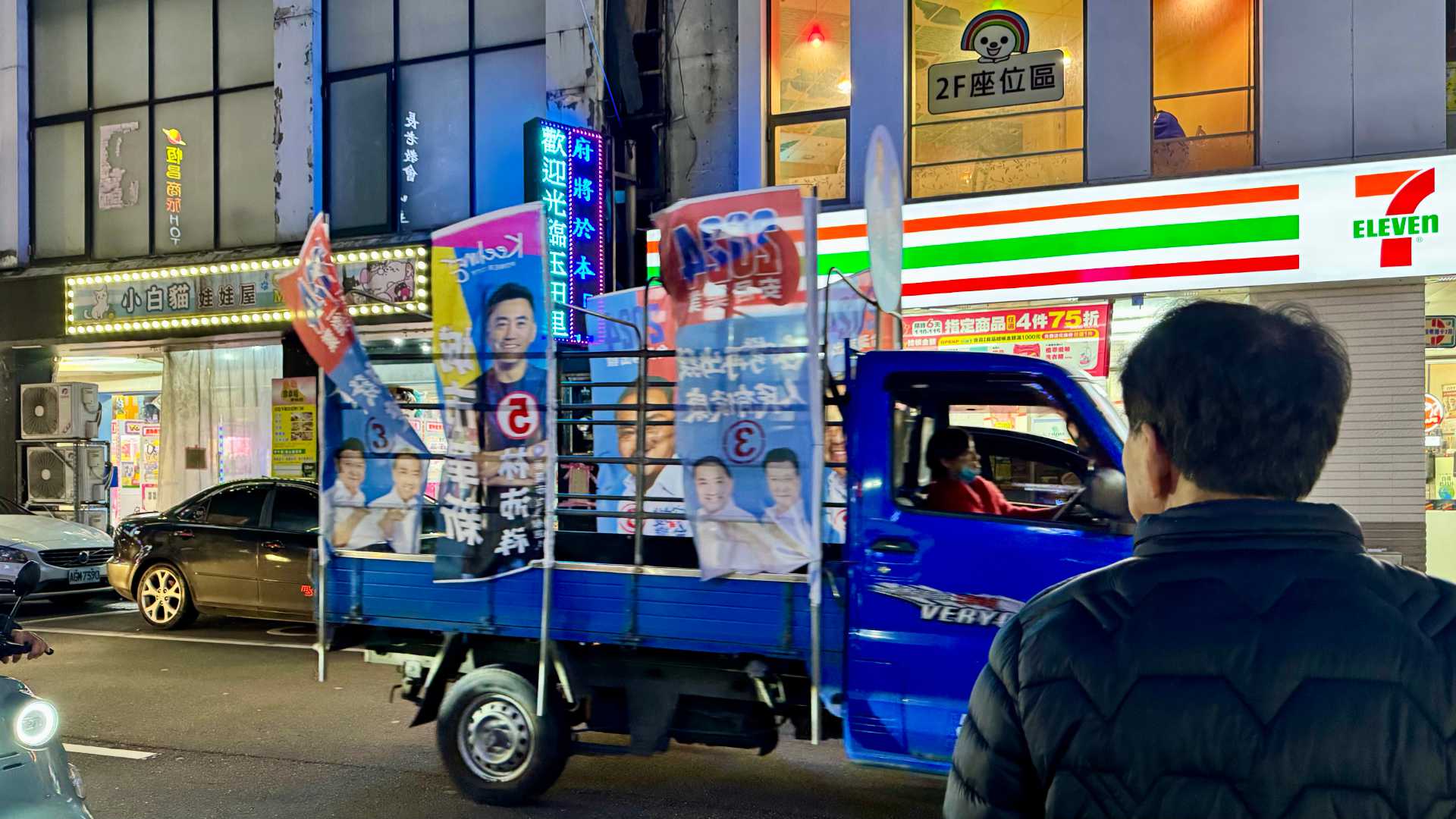 A small truck driving past pedestrians, with political flags and banners on the back.
