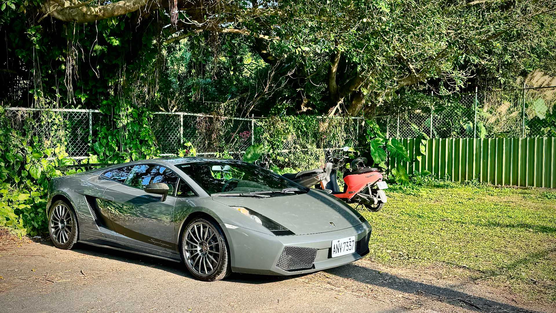 A Lamborghini Gallardo at one end of a paved parking lot, next to two scooters parked on the grass.