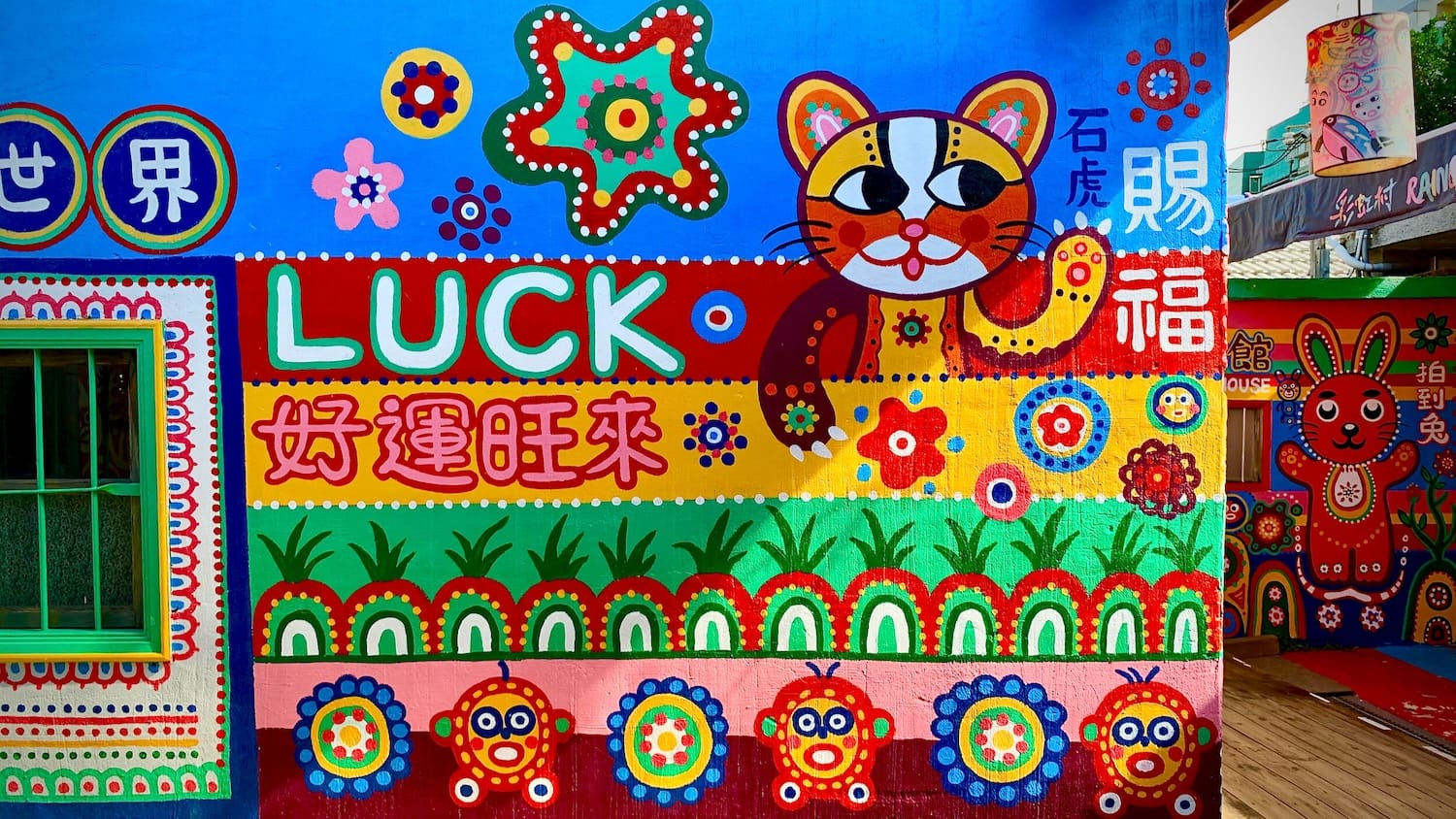 A Taiwan Leopard Cat mascot painted on the wall of a house in Rainbow Village. Next to the cat, the word LUCK is painted on the wall.