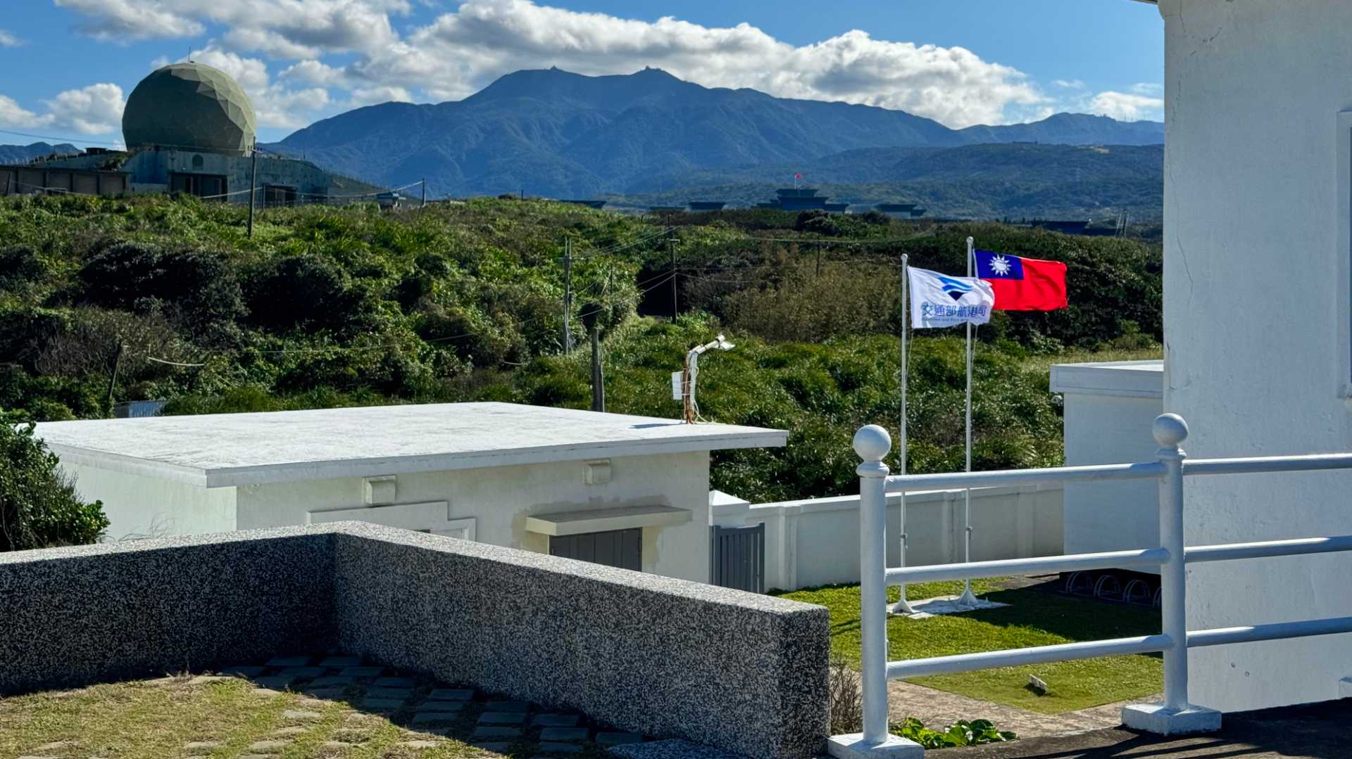In the foreground, some white buildings and a Taiwan flag. Across vegetation in the distance is a radar dome and other military buildings.