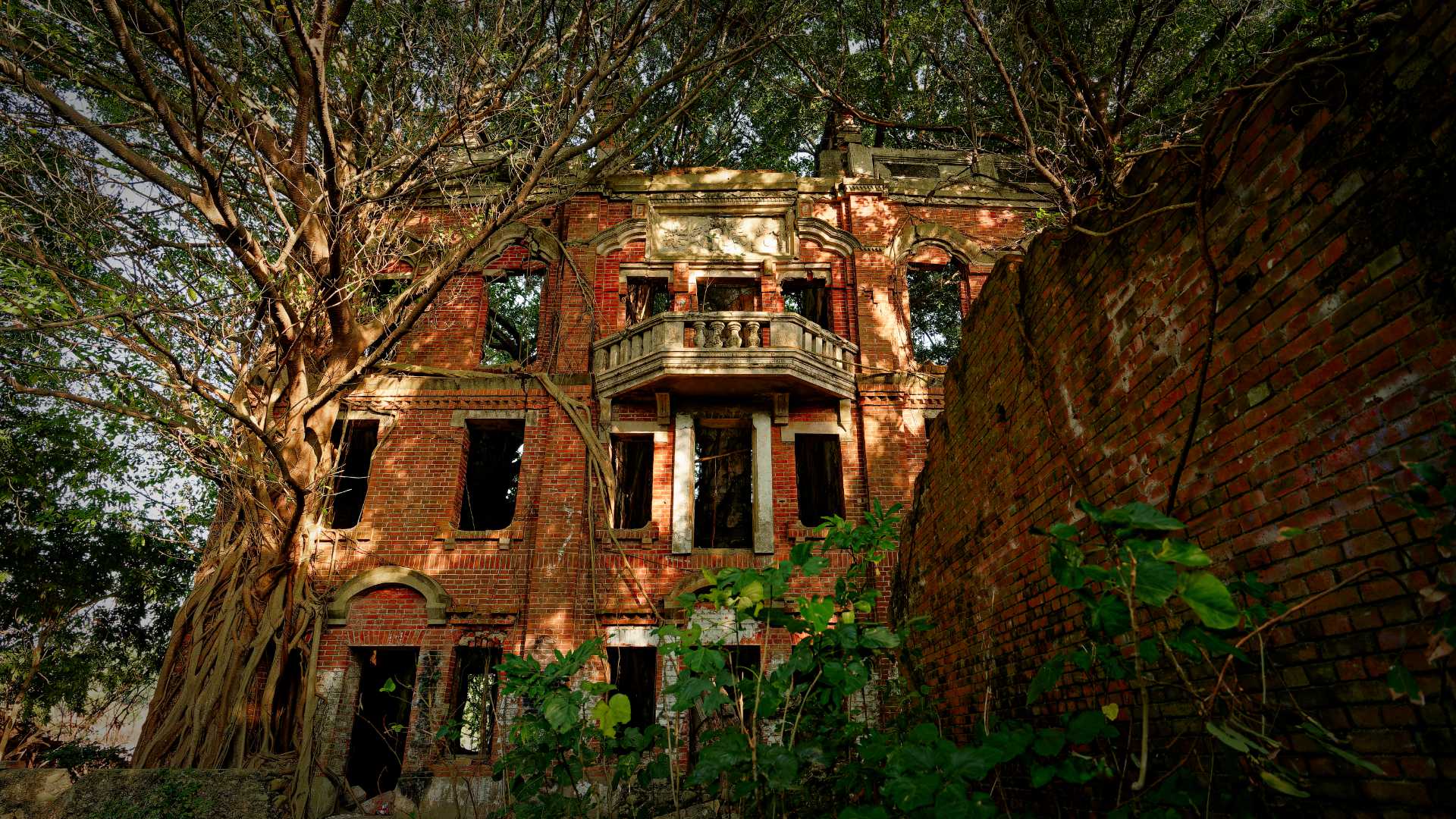 The front of Minxiong Haunted House, an abandoned three-story brick mansion a Chiayi forest in Taiwan. The windows and doors are missing. There are tall trees growing out from the masonry, all across the building.