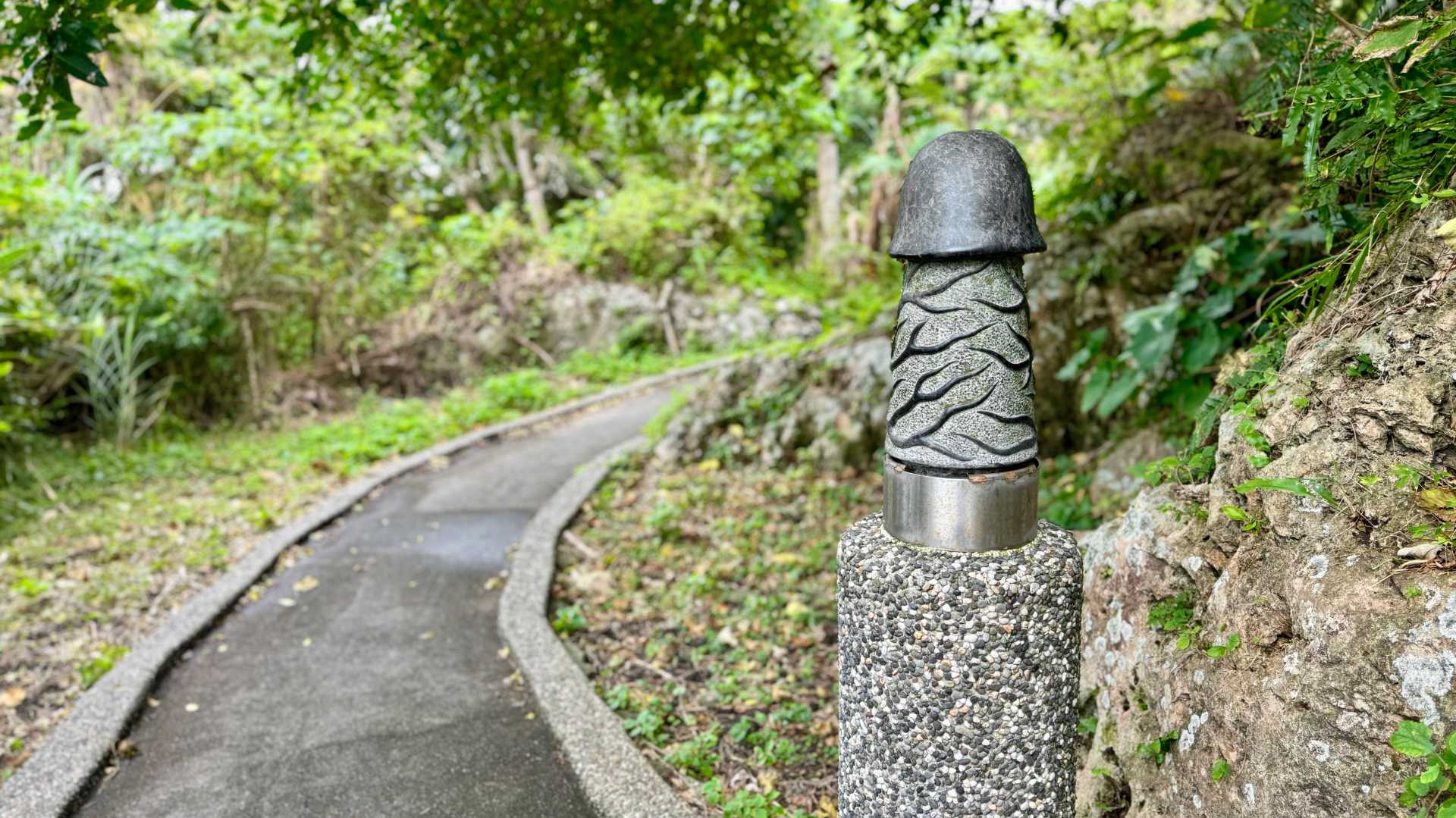 A small stone sculpture of an erect penis, perhaps 30cm tall, mounted on a stone post next to a curved pathway.