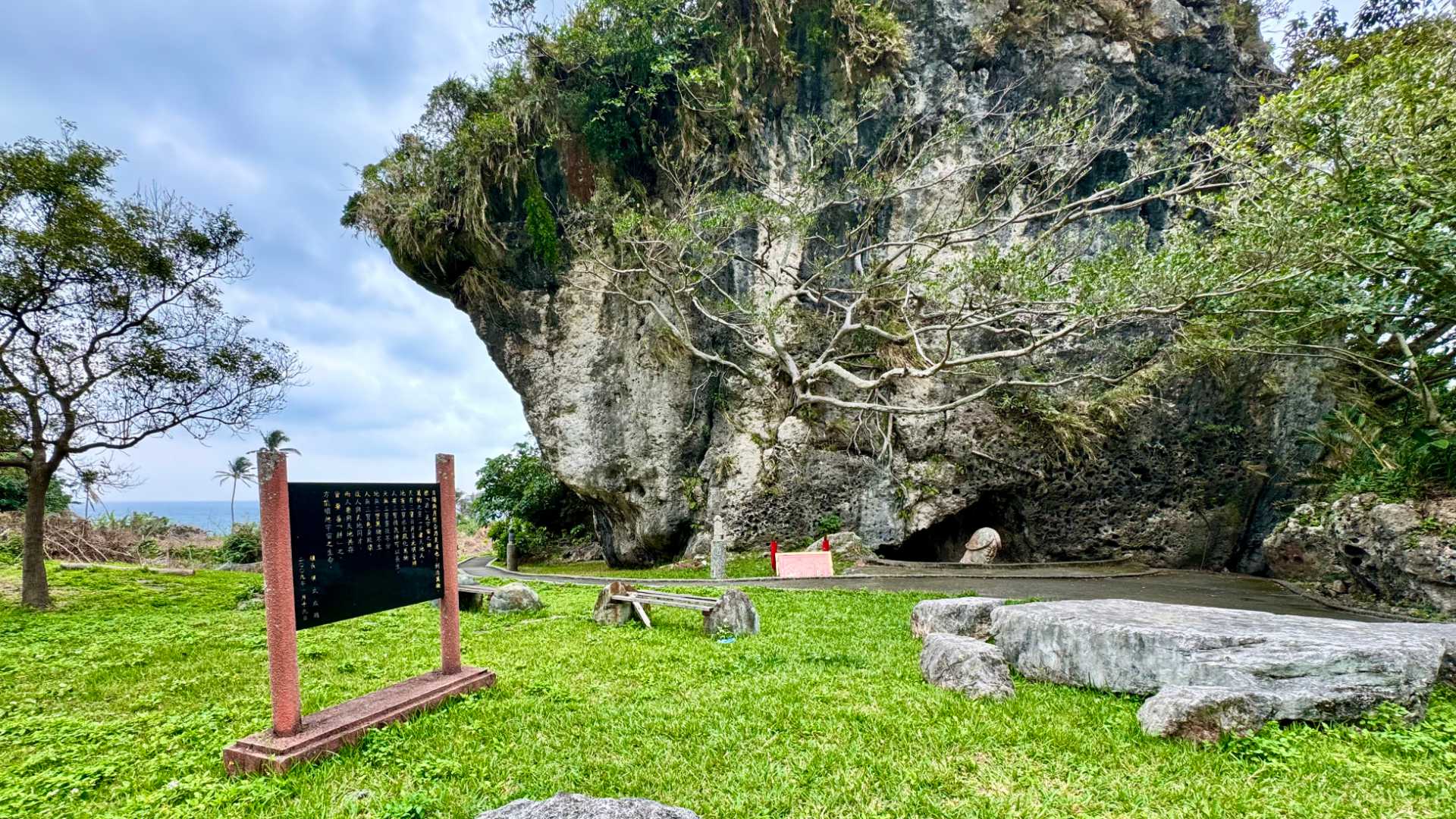 A grassy area with some wooden seating and a cave visible a few meters away. There is something poking out of the cave.