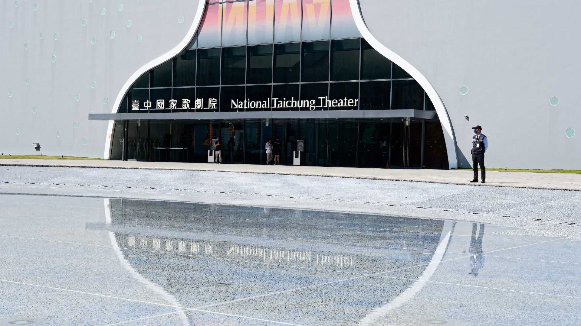 A security guard at the entrance to National Taichung Theater, reflected in a shallow pool of water.