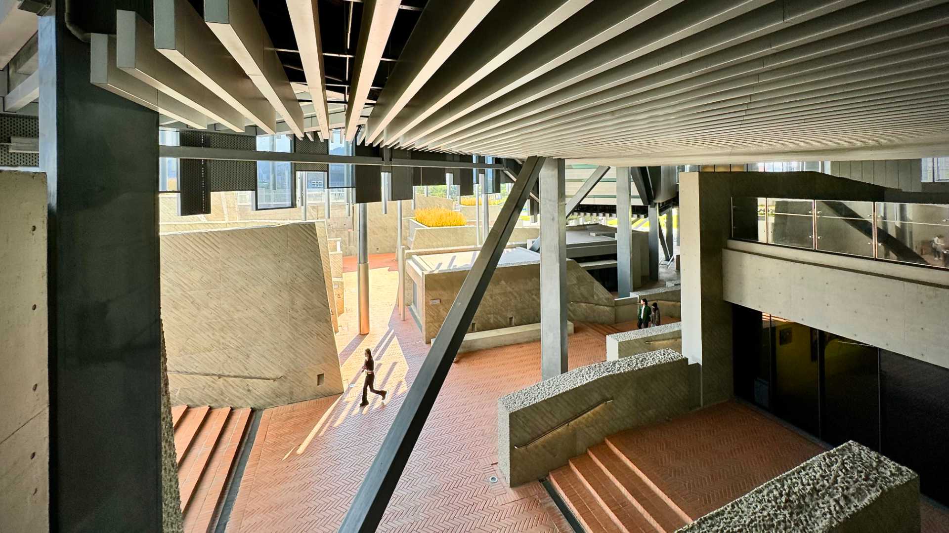 A double-height space beneath a building, open to the outdoors, comprising multiple levels of tiled spaces with low concrete walls.