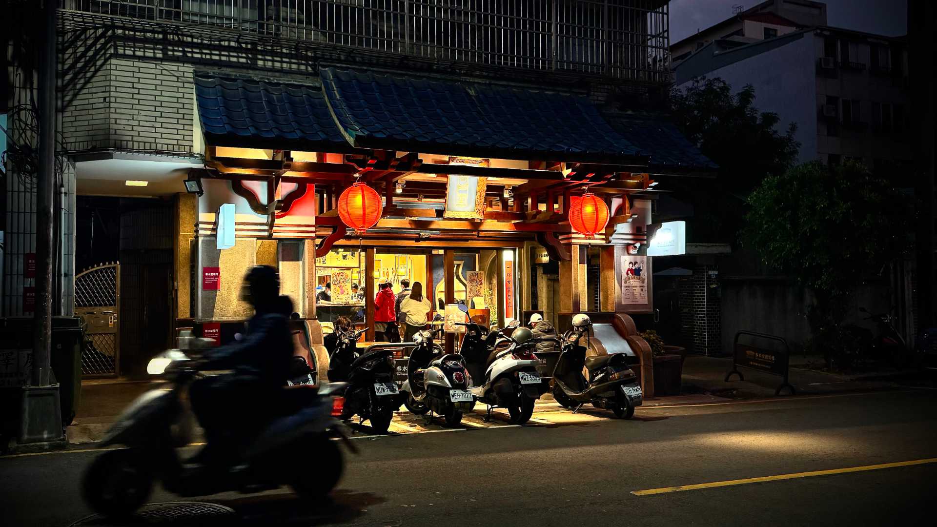 Exterior of the famous Chun Shui Tang teahouse. It's nighttime. There are scooters parked in a row outside the shop, which features a tiled roof and red lanterns.