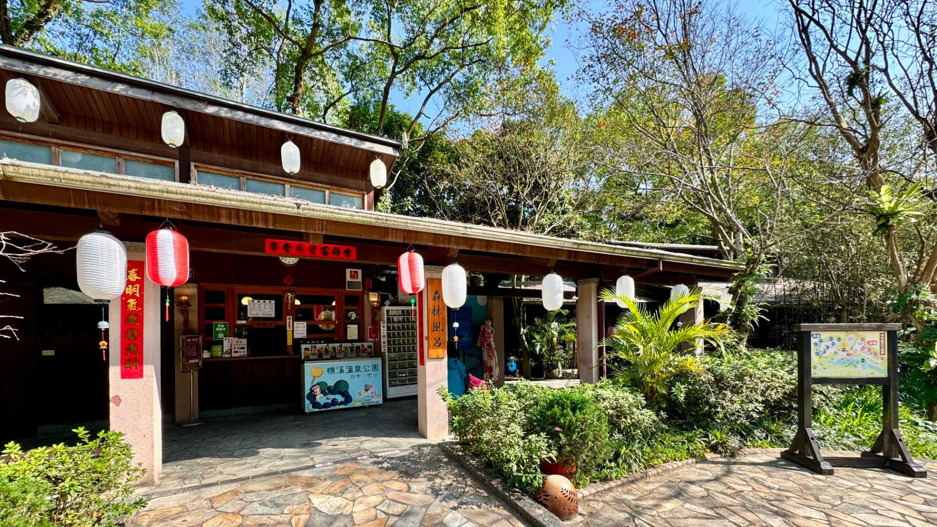 A Japanese-style wooden building with paper lanterns hanging out the front, set in a leafy green park.