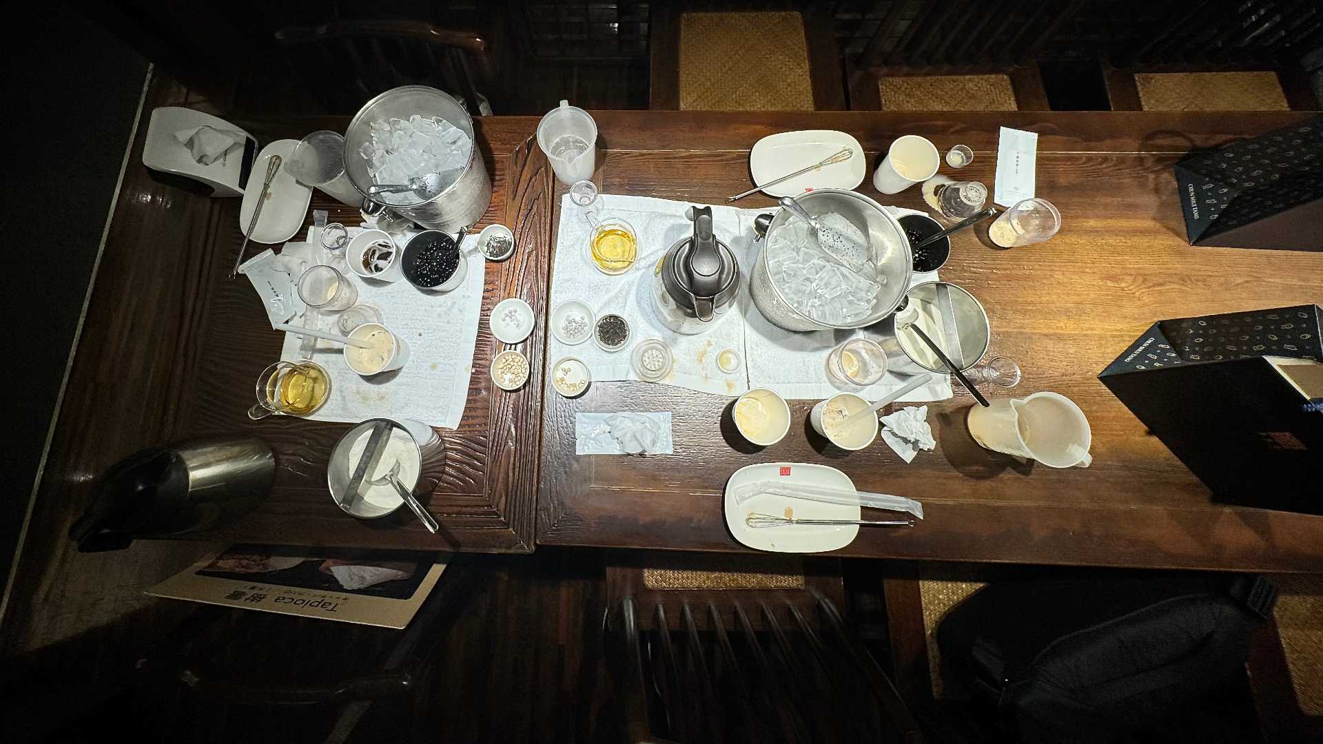 A messy tabletop covered in paper cups, kitchen utensils, and bubble tea ingredients.