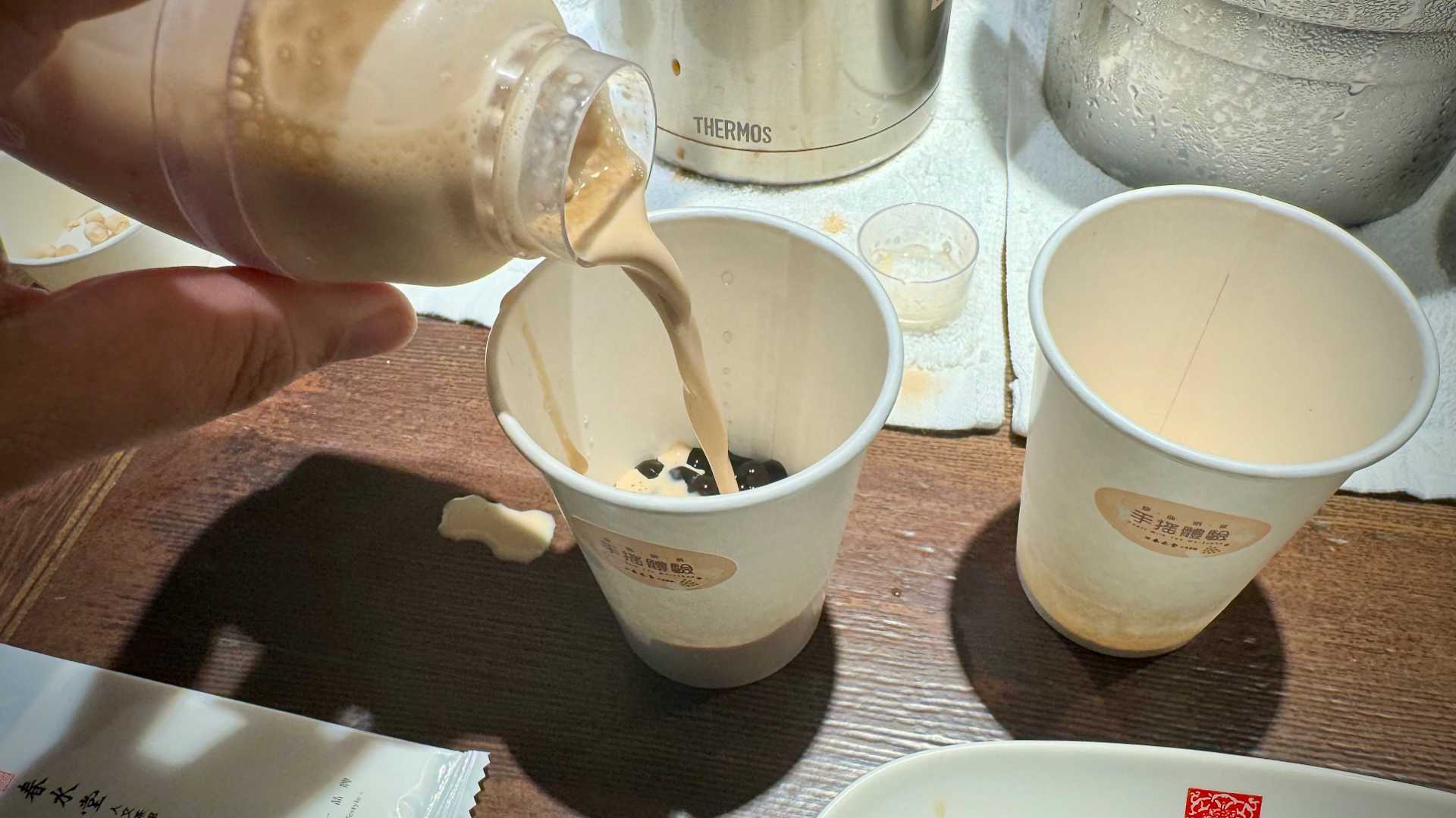 Pouring shaken milk tea from a cocktail shaker into a paper cup of tapioca balls.
