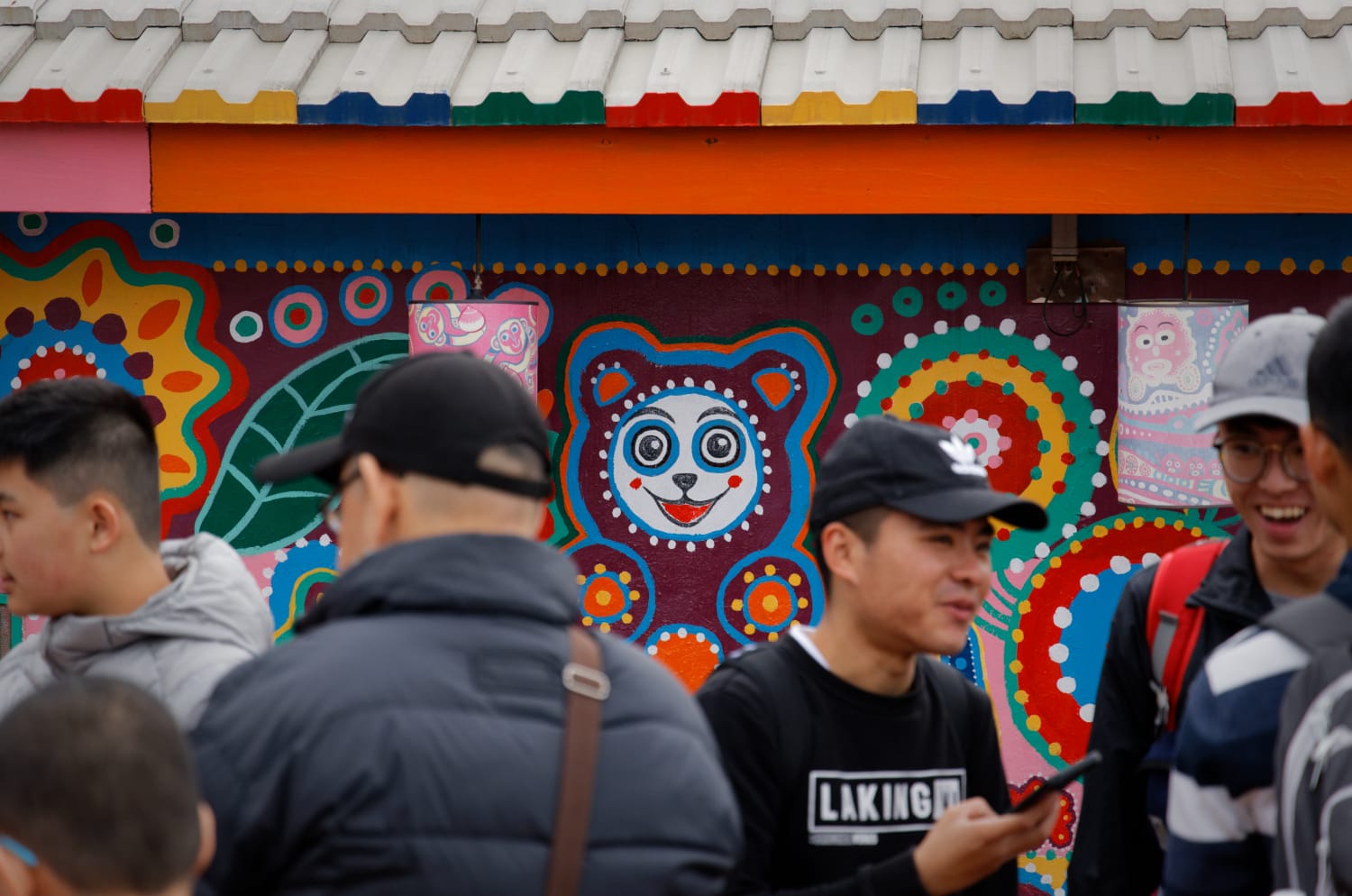 People talking in front of a mural that covers the wall of a house of Rainbow Village, featuring a cartoonish happy bear.