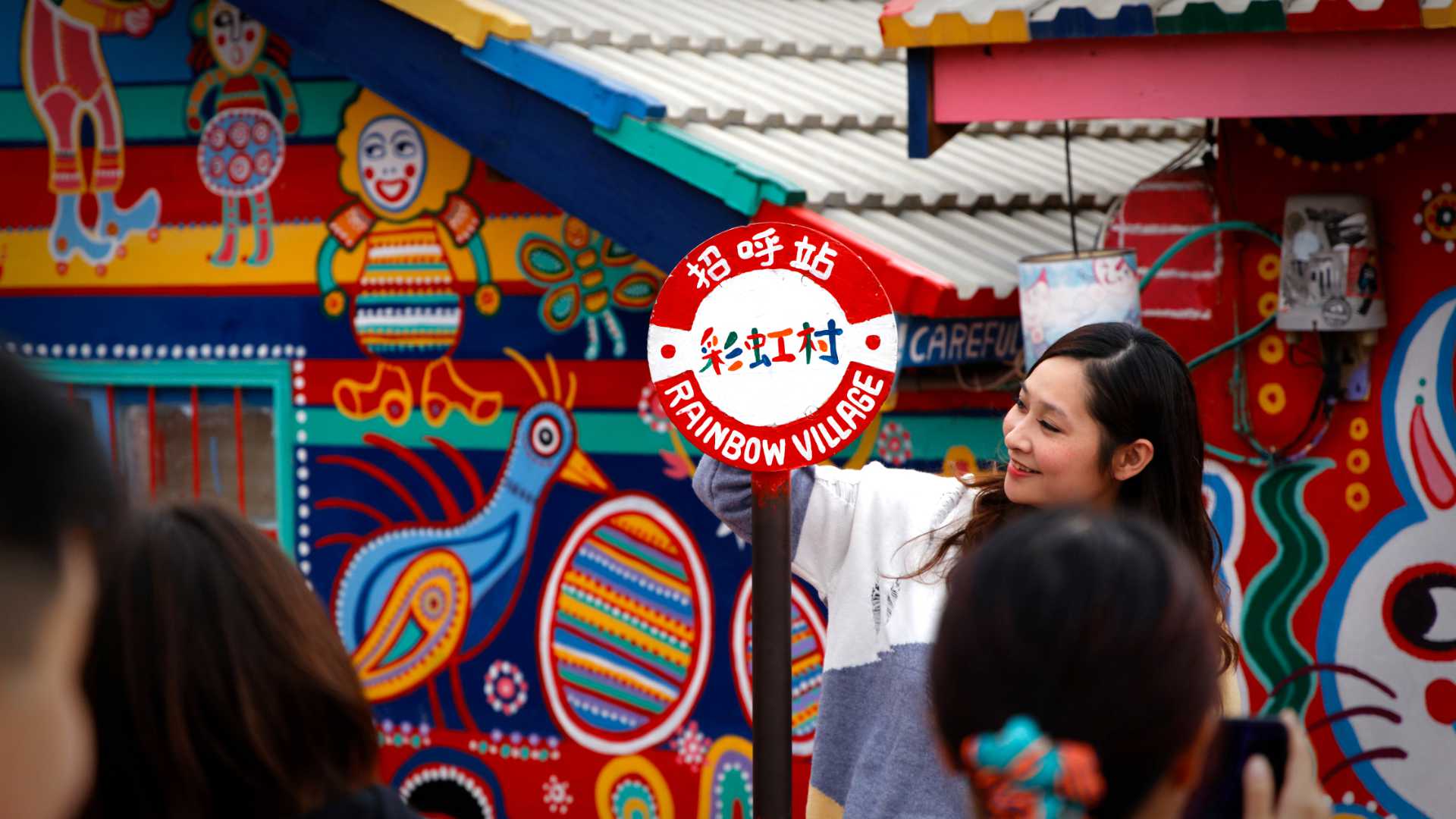 A happy young woman looking at a sign that says Rainbow Village. Brightly-painted village buildings are visible behind her, with crowds of people in the foreground.