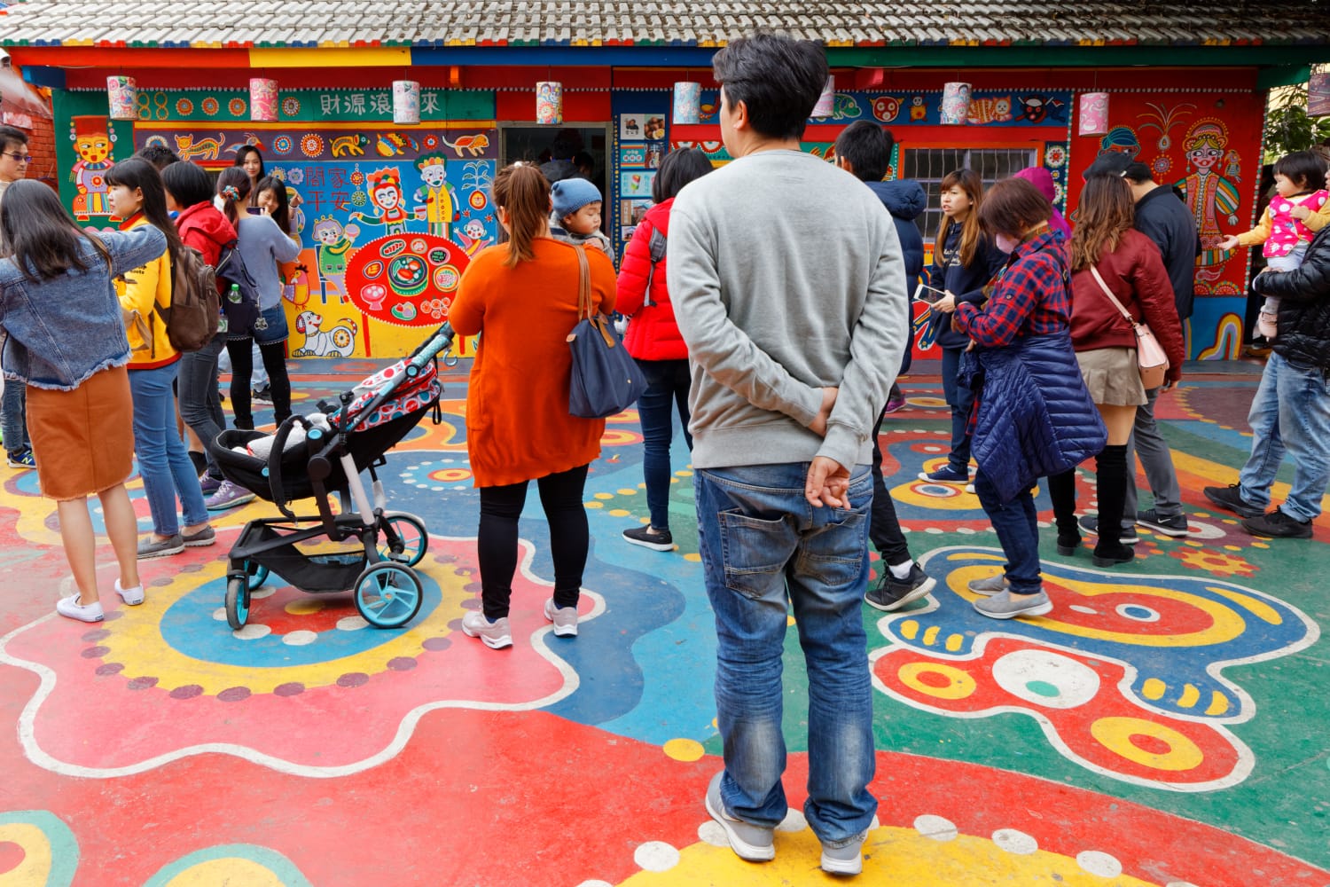 The forecourt outside Rainbow Village in Taichung, Taiwan. The concrete ground is covered in colorful art, as are the buildings in the distance. The forecourt is crowded with people.