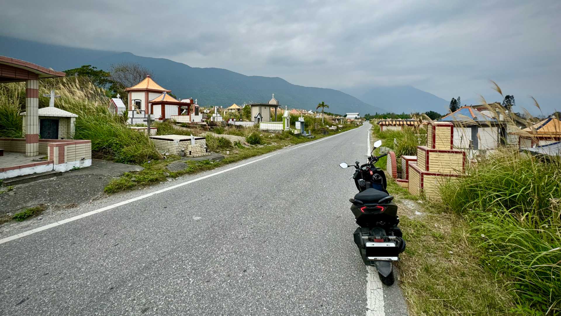 A motorcycle parked on the side of a one-lane road through the middle of a grassy cemetery.