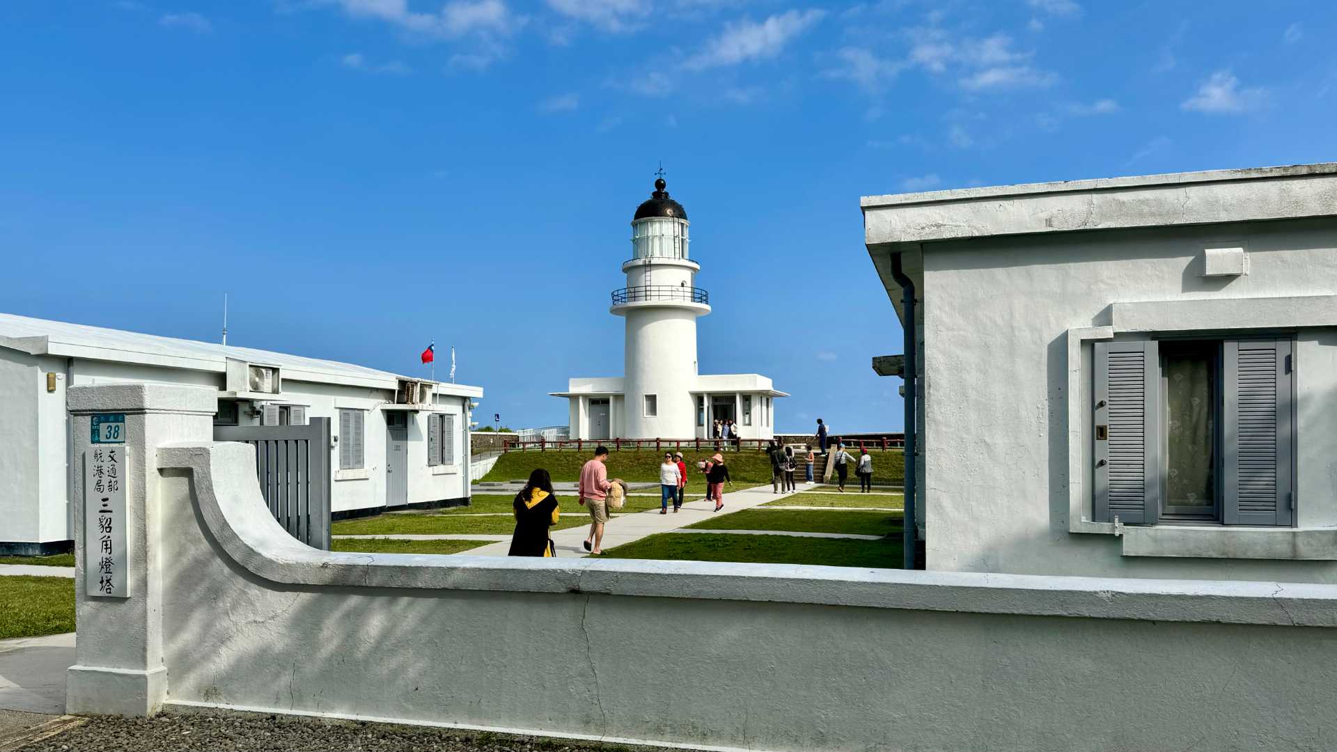 Santiao Chiao Lighthouse, a white lighthouse on immacuately-maintained grounds. Single-story while buildings, and a Taiwan flag, line the sides of the lighthouse grounds. There are two-dozen tourists wandering around. It looks like a warm, sunny day.