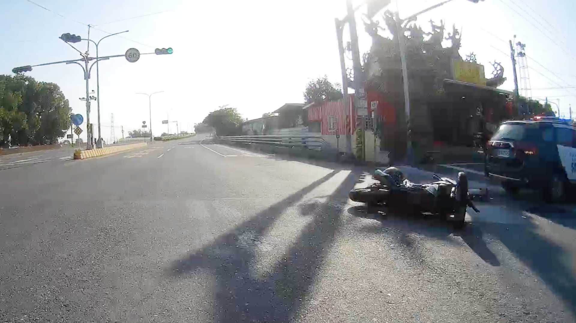 Dashcam screenshot of a scooter fallen on its side in the middle of an intersection. A police car with flashing lights is parked next to it. There is a helmet in the scene but it’s unclear if a person is laying there or not.