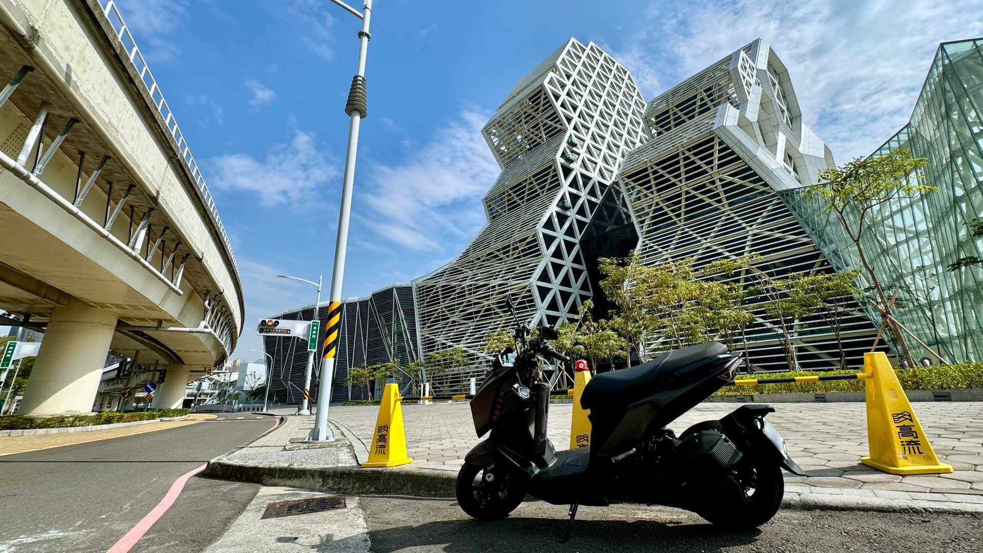 A scooter parked on the street outside Kaohsiung Music Center. A short, lightweight, yellow-and-black barrier is erected on the other side of the scooter.