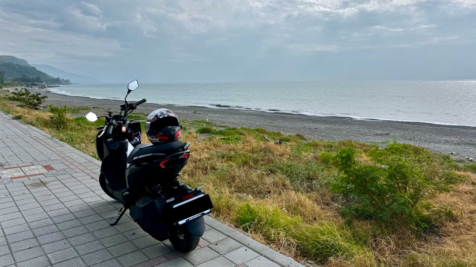 A scooter parked on a tiled path next to a stony beach and a calm sea.