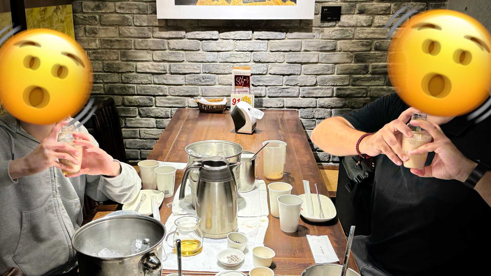 Two people sitting either side of a table covered in bubble tea ingredients, shaking cocktail shakers.