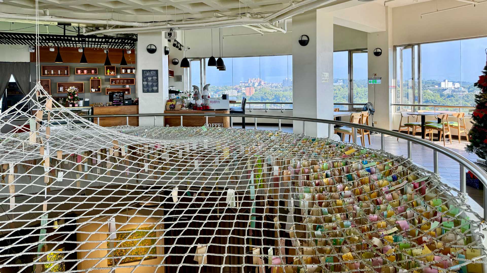 The cafe on level 11 of Sun-Shooting Tower in Chiayi. The city can be seen through large windows, and in the foreground a void space has been covered by a net, off which are hanging hundreds of colorful cards with wishes written on them.