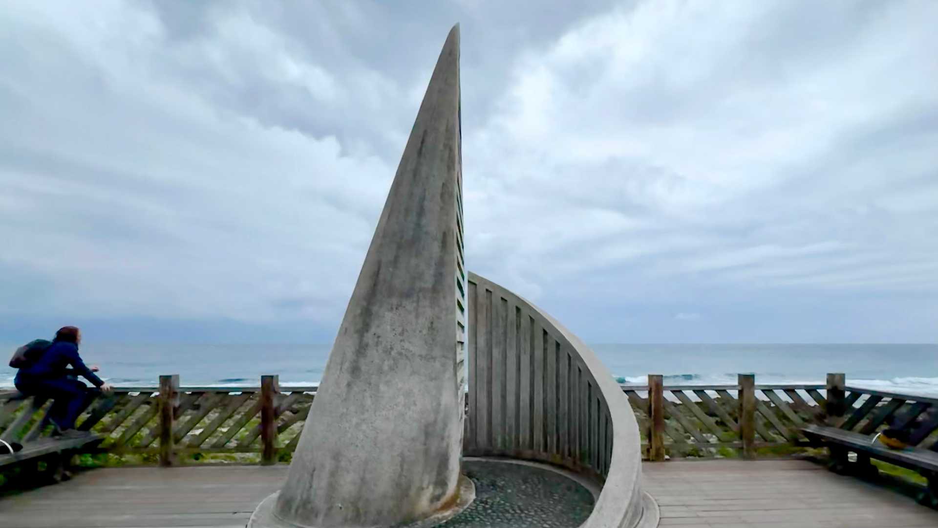 A spiral concrete sculpture on a wooden viewing platform. One person is sitting on the handrailing. The ocean is in the distance.
