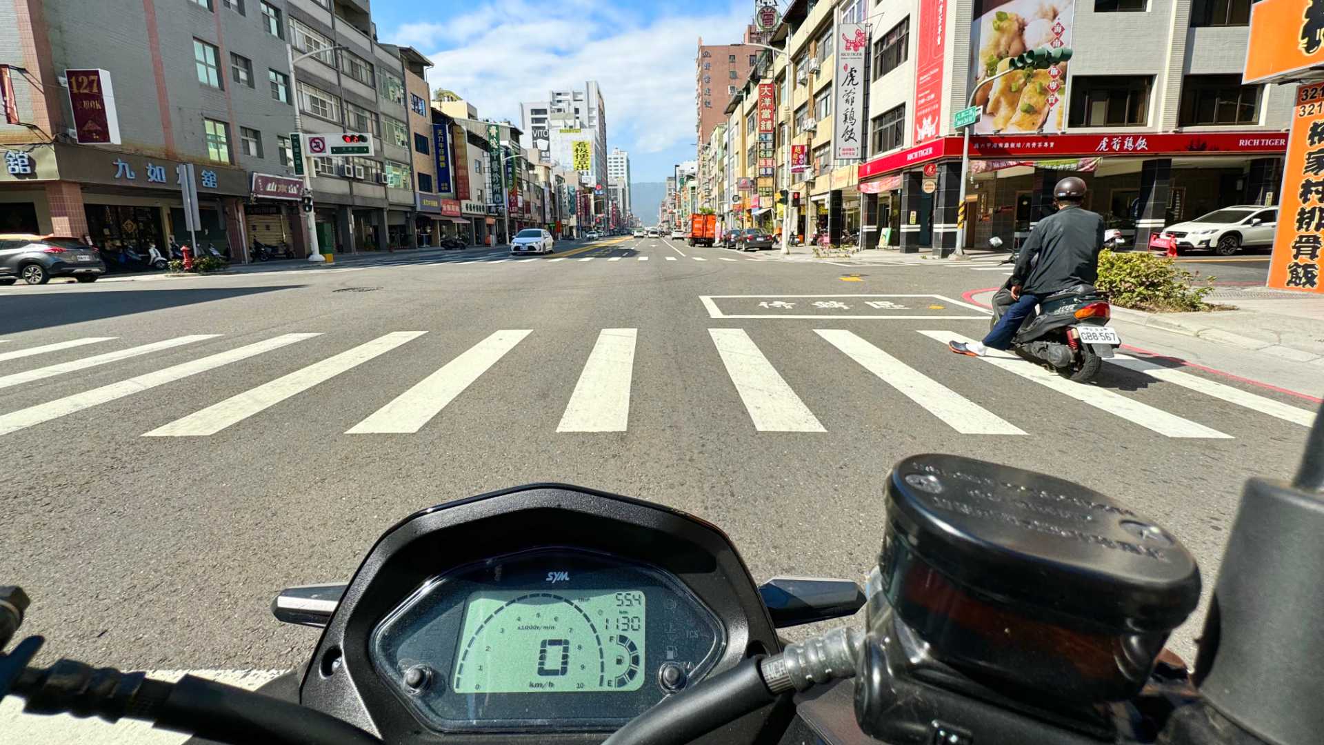 A point-of-view shot taken from a motorcycle while stopped at a red light, of a four-lane road in Kaohsiung with Monkey Mountain barely visible in the far distance.