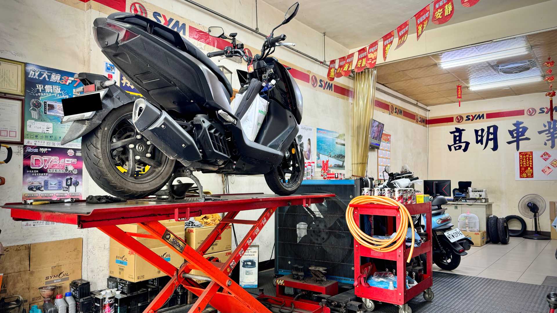 An SYM MMBCU scooter on a hoist in a mechanic workshop.