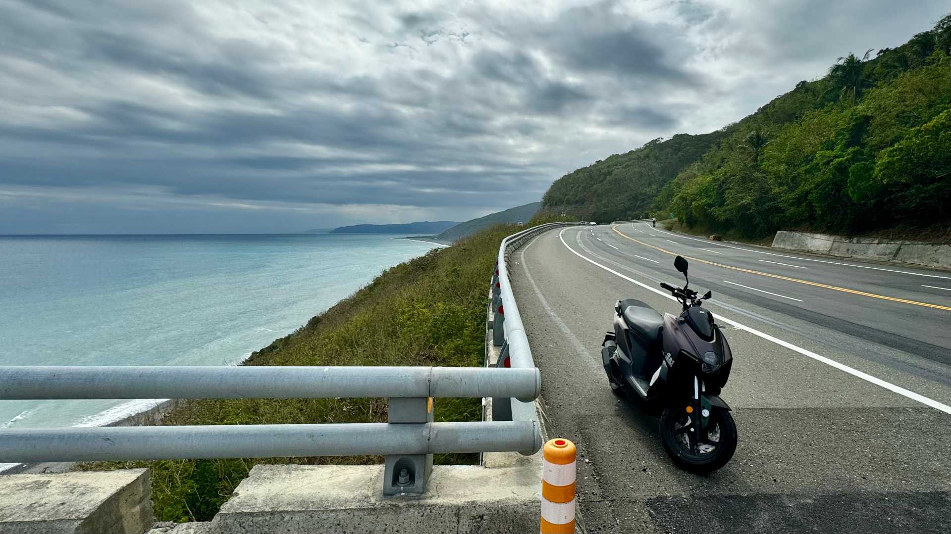 A scooter parked on the side of a four-lane coastal road, nested between forested mountains and a stony beach.