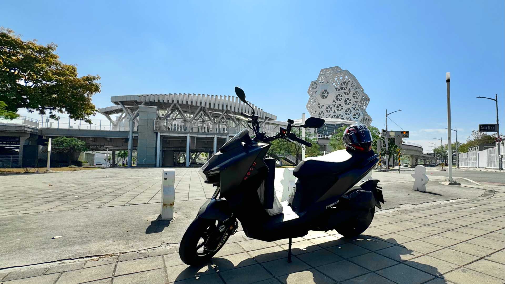 An SYM MMBCY scooter parked outside Kaohsiung Music Center. A motorcycle helmet rests on the scooter seat. It’s a beautiful sunny day.
